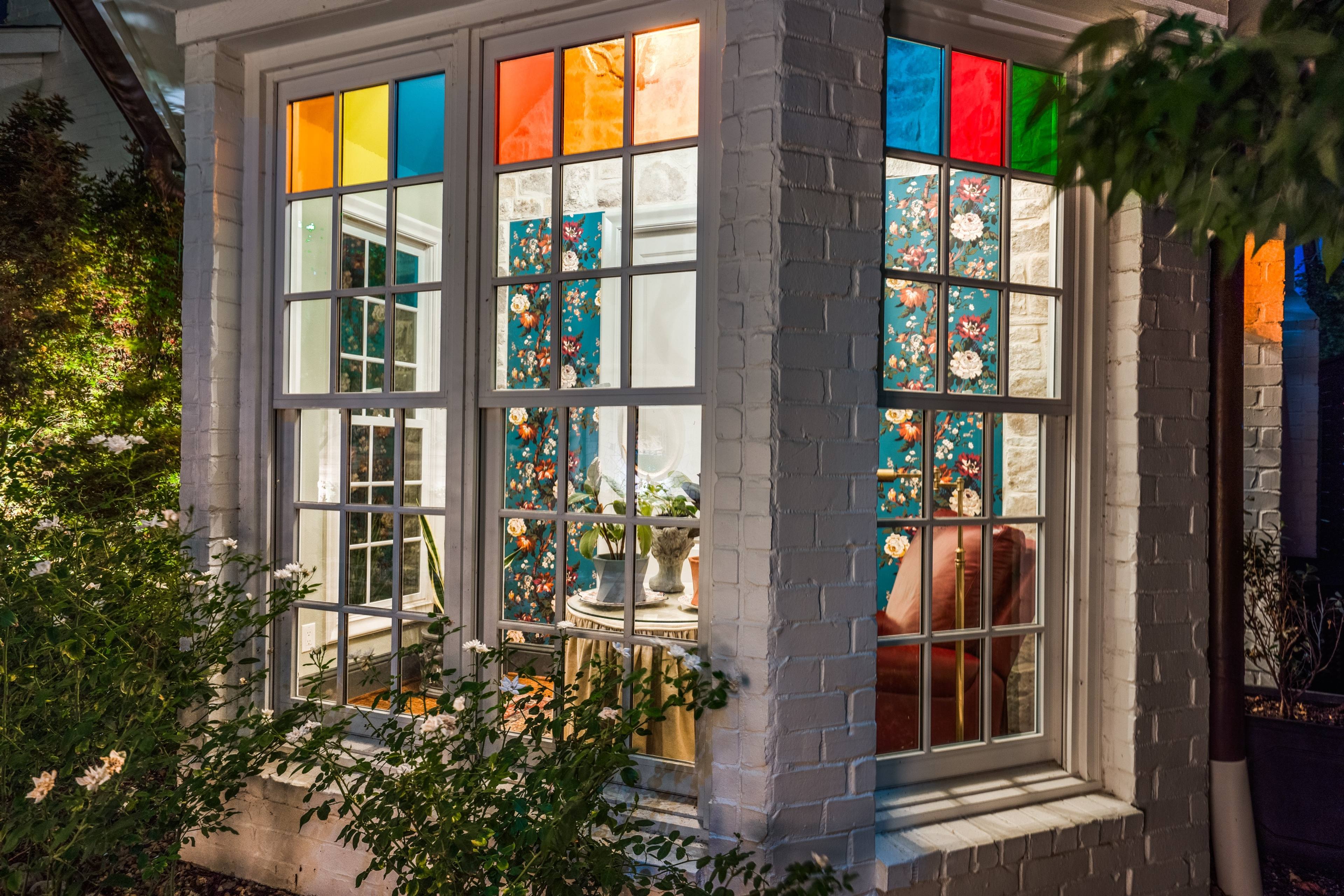 Evening twilight view of a painted brick bay window with colorful stained glass panels framing a floral wallpapered breakfast nook at a historic Lakehurst estate.