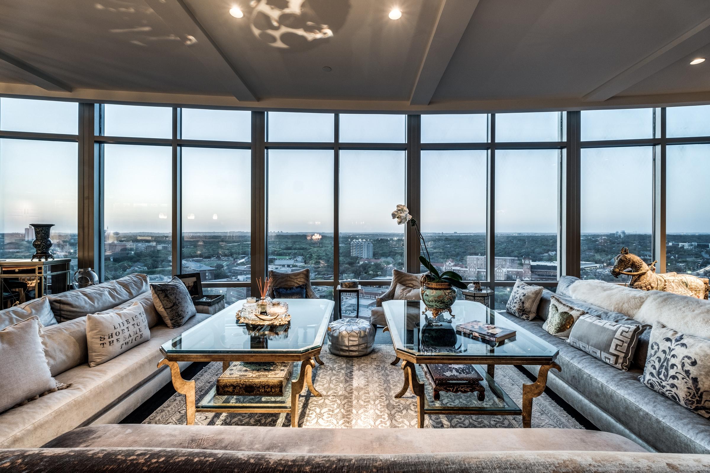 Luxury high-rise living room with facing sofas, glass coffee tables, and floor-to-ceiling windows over San Antonio at dusk