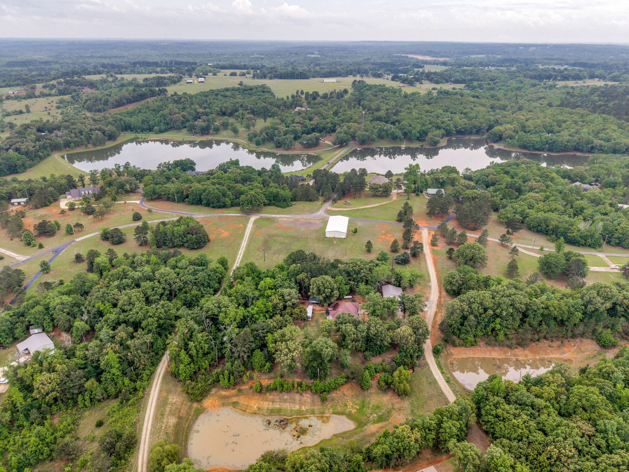 Wide aerial of a rural Lindale, TX lakefront property — main home, outbuilding, curving drive, pond, and sprawling East Texas tree cover (before Aerial Green Grass correction).