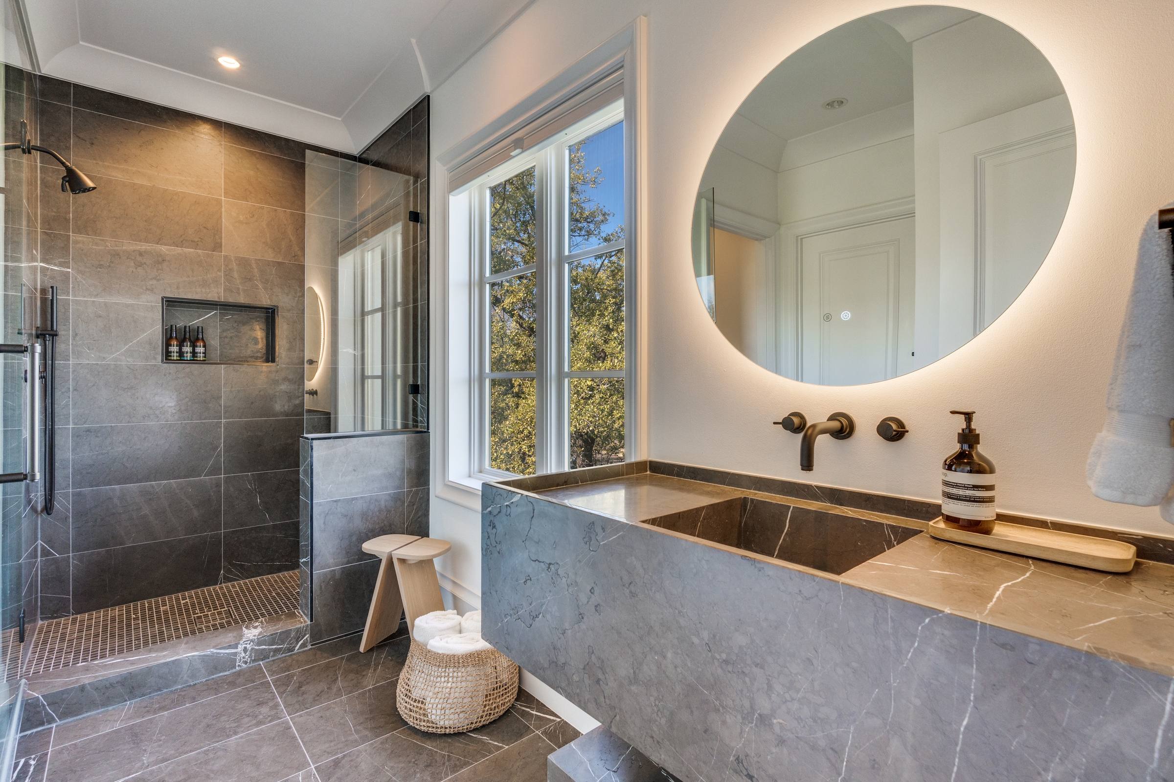 Modern bathroom with dark marble vanity, backlit round mirror, and walk-in shower in Highland Park, Texas