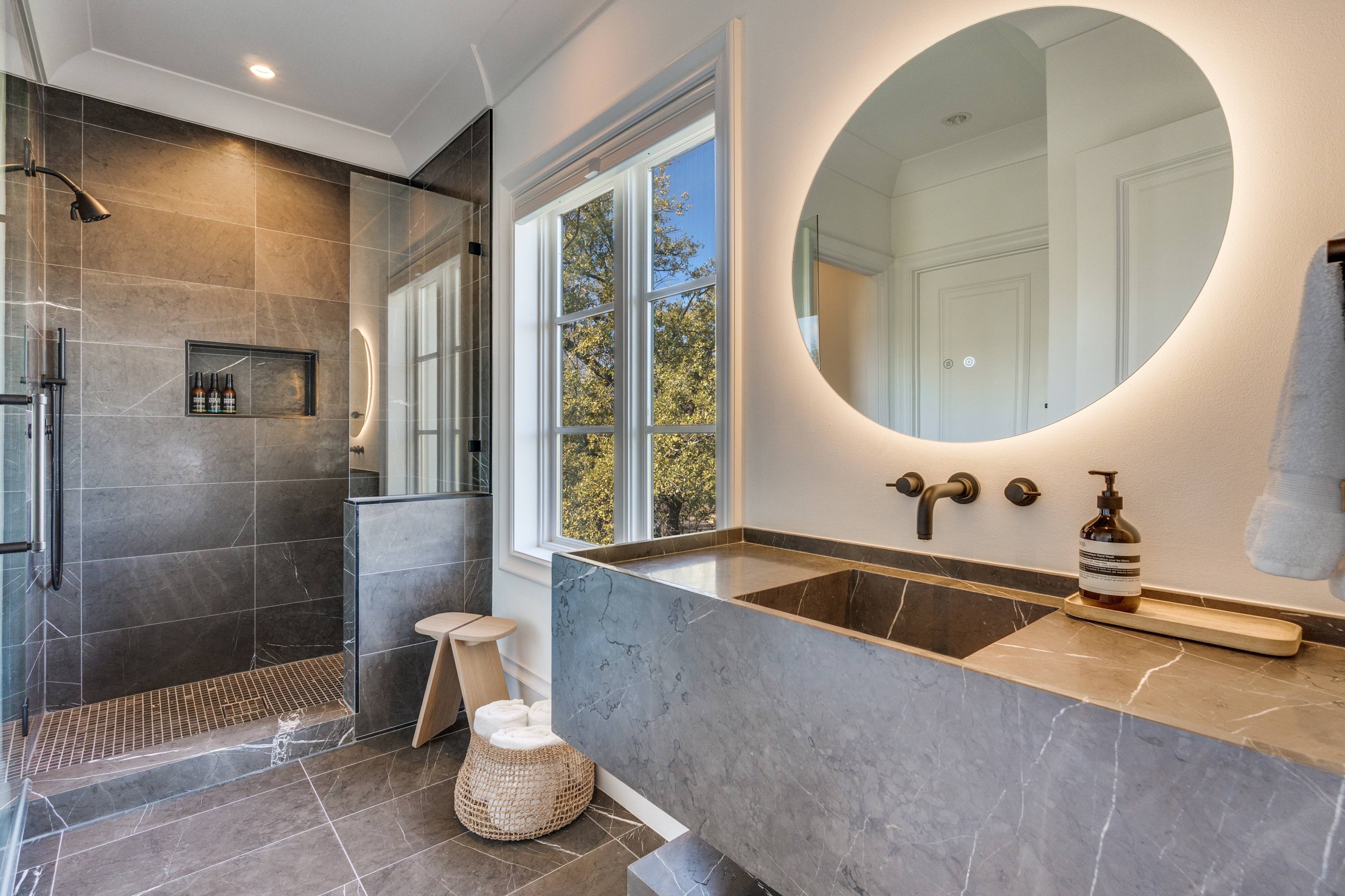 Modern bathroom with dark marble vanity, backlit round mirror, and walk-in shower in Highland Park, Texas