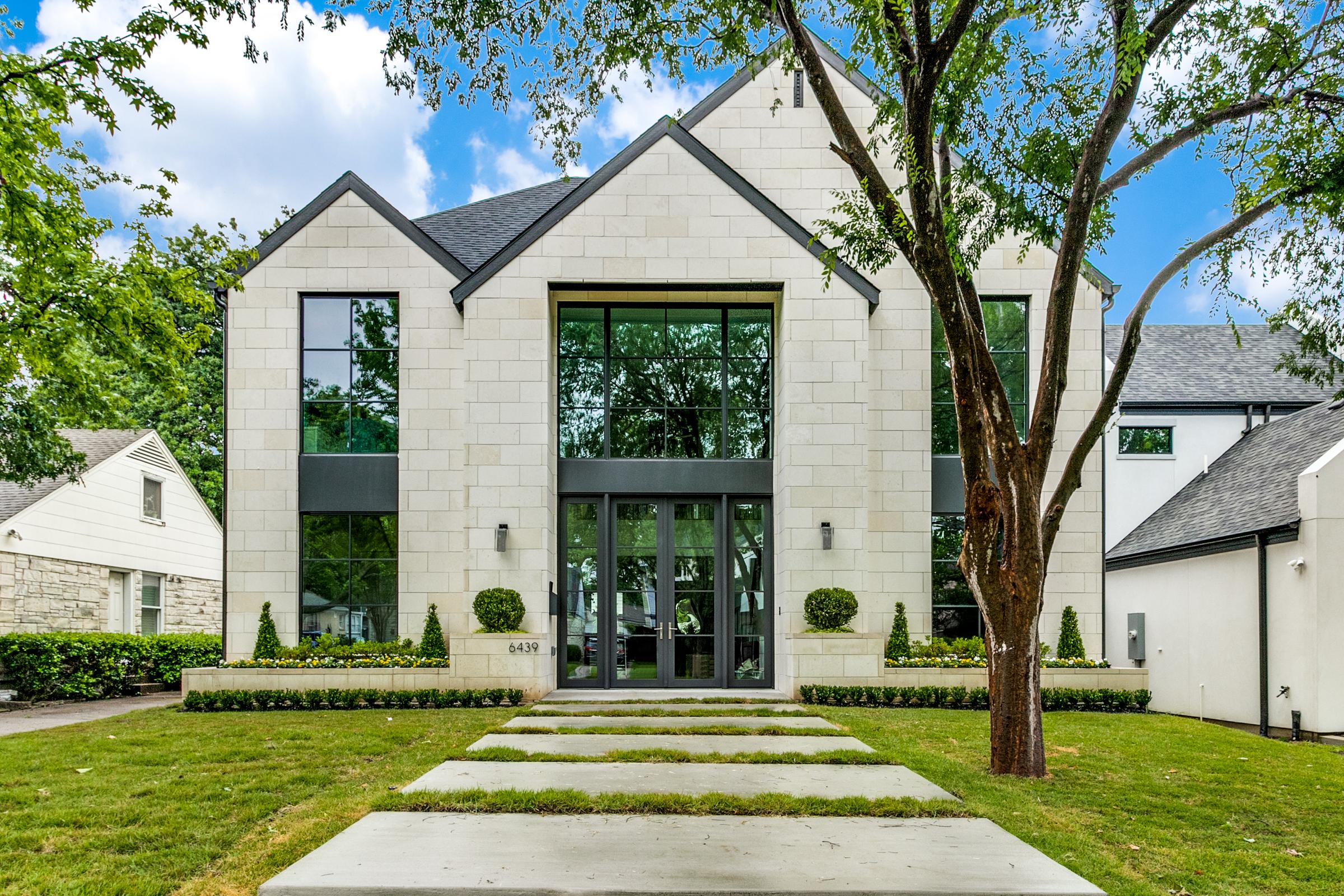 Modern luxury home in Dallas, TX with three symmetric gables, light stone exterior, and a dramatic floor-to-ceiling wall of windows over a steel-framed front entry