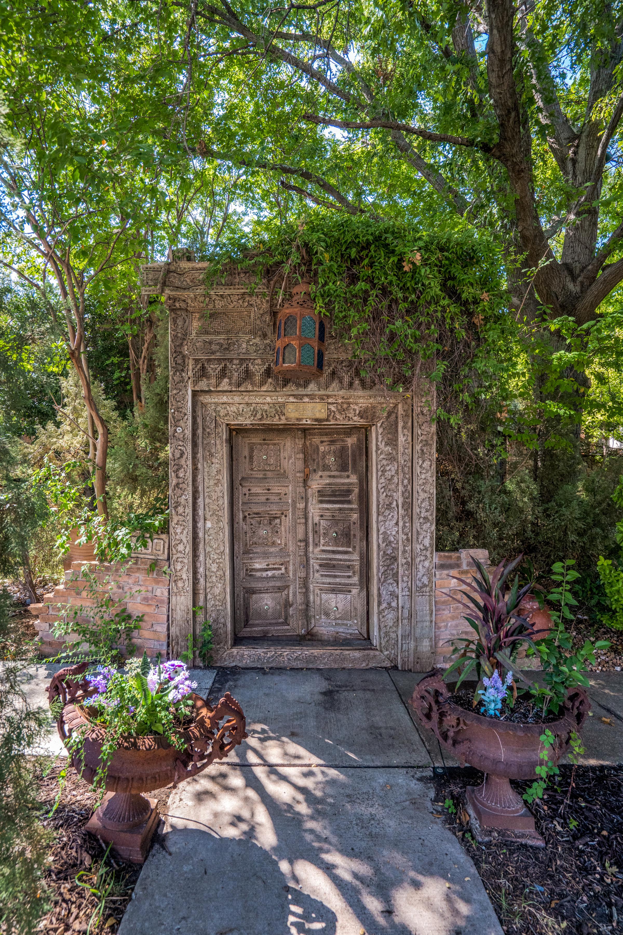 Ornately carved antique wood door set into a stone archway with vines, a vintage lantern, and symmetrical urn planters on a Plano, TX garden walkway.