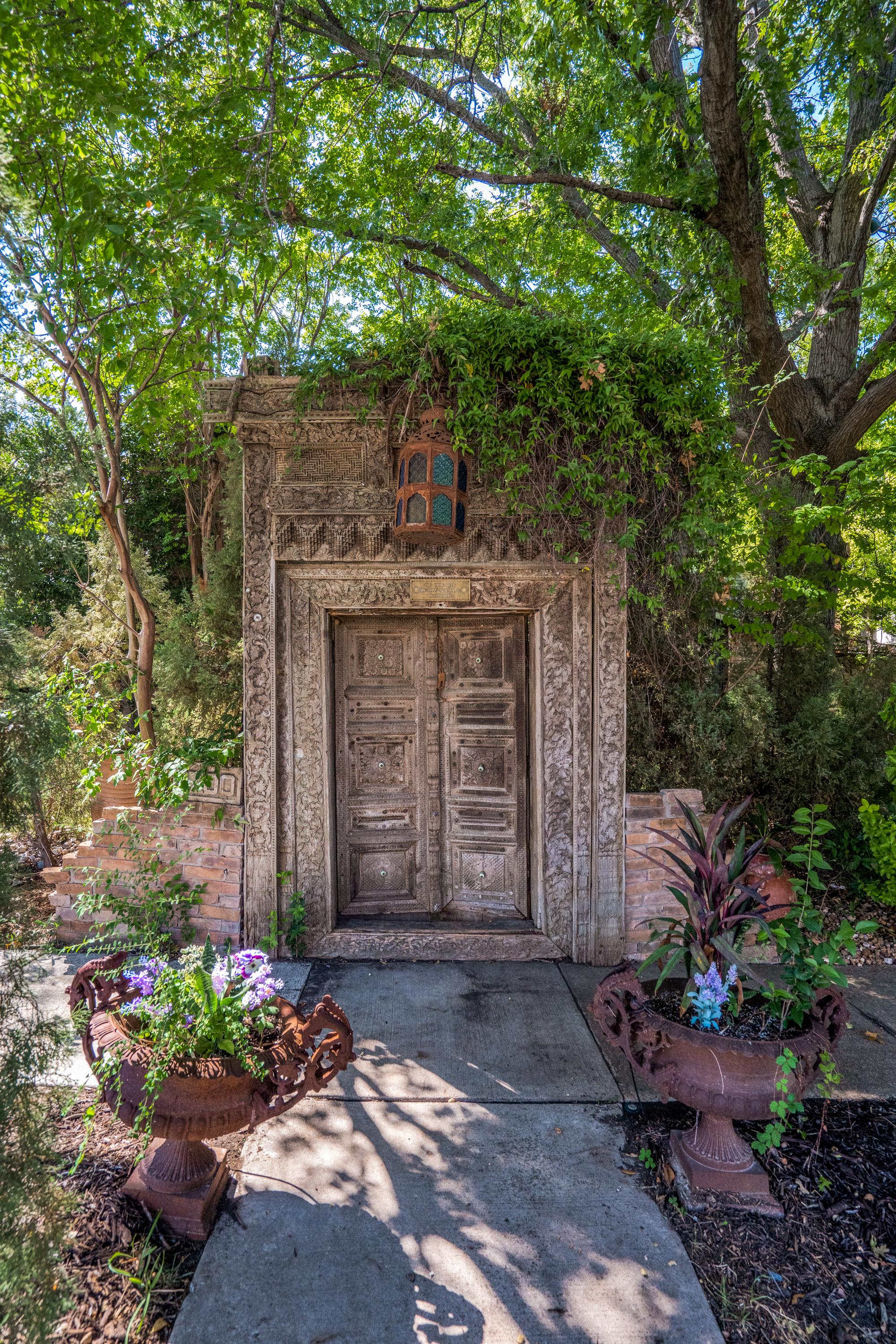 Ornately carved antique wood door set into a stone archway with vines, a vintage lantern, and symmetrical urn planters on a Plano, TX garden walkway.