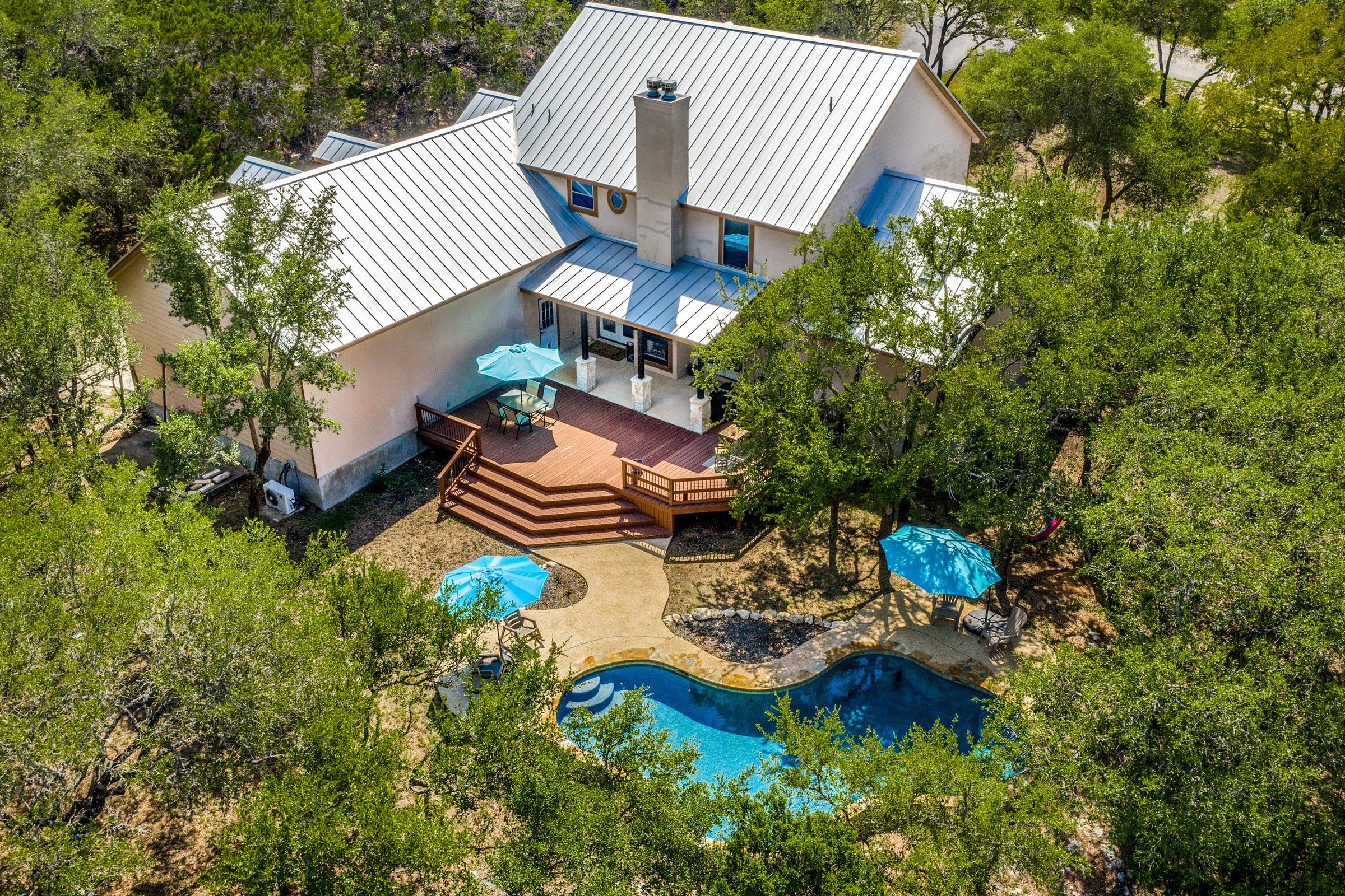Aerial drone photograph of a luxury Hill Country home with metal roof, multi-level wood deck, freeform pool with rock waterfall, and mature live oaks in Spring Branch, Texas