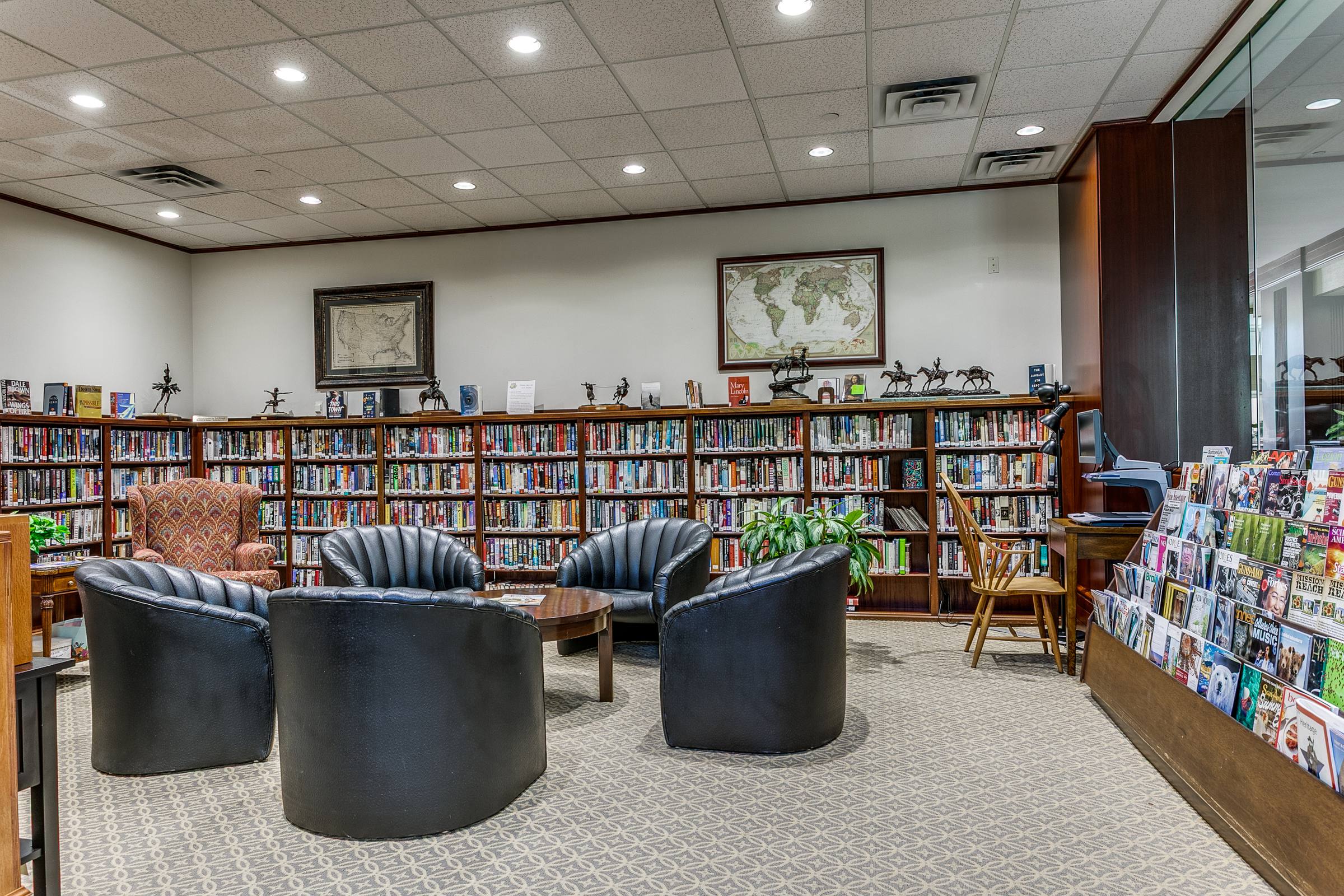 San Antonio, TX community library interior with wood bookshelves, black leather club chairs around a small table, framed maps, equestrian bronzes, and a magazine rack — a clubhouse-style reading lounge.