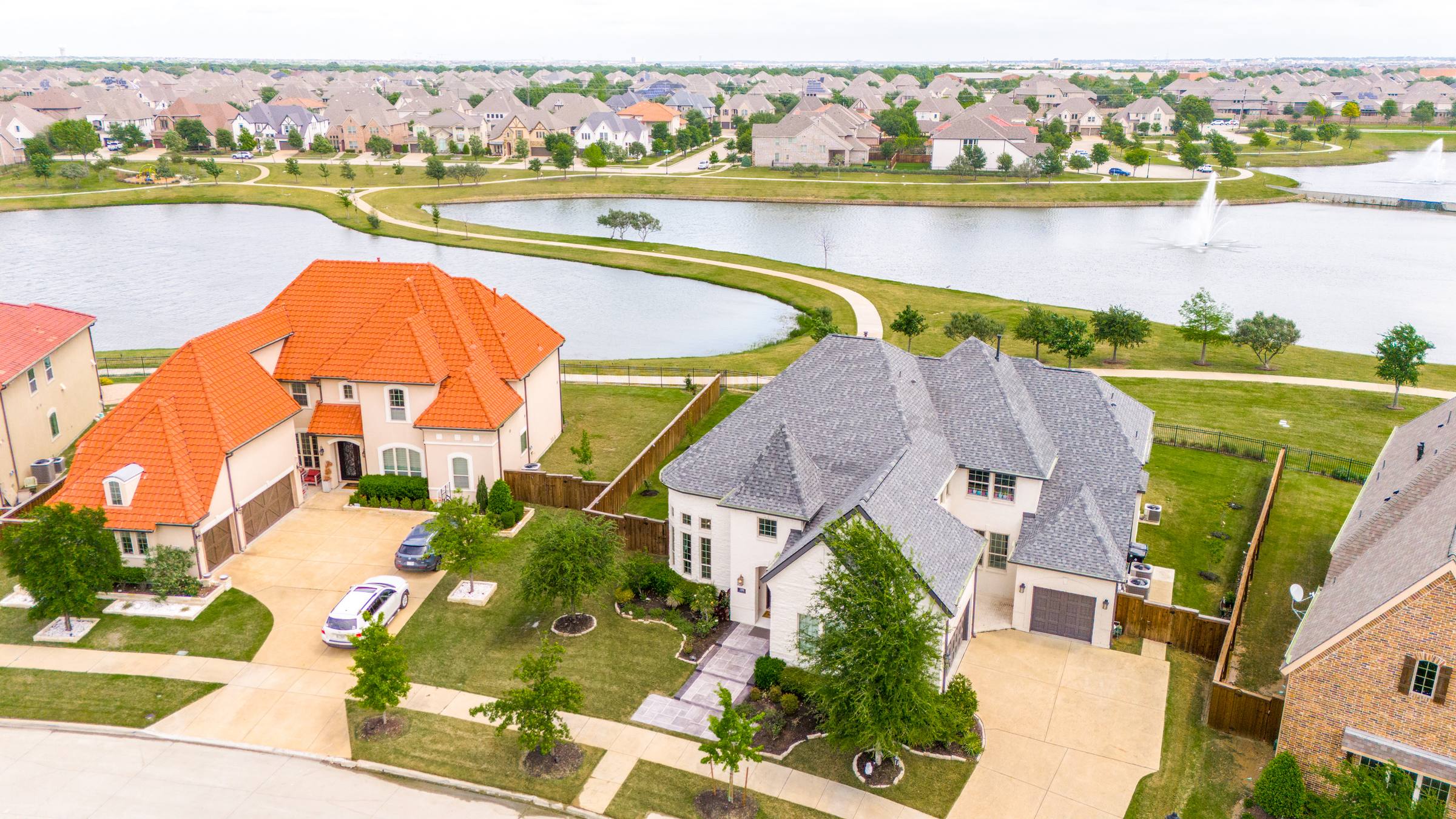 Aerial of a Frisco, TX lakeside suburban street — two custom homes in the foreground flanking a shared cul-de-sac, a walking path around the pond, and rooftops stretching to the horizon (before Aerial Green Grass correction).