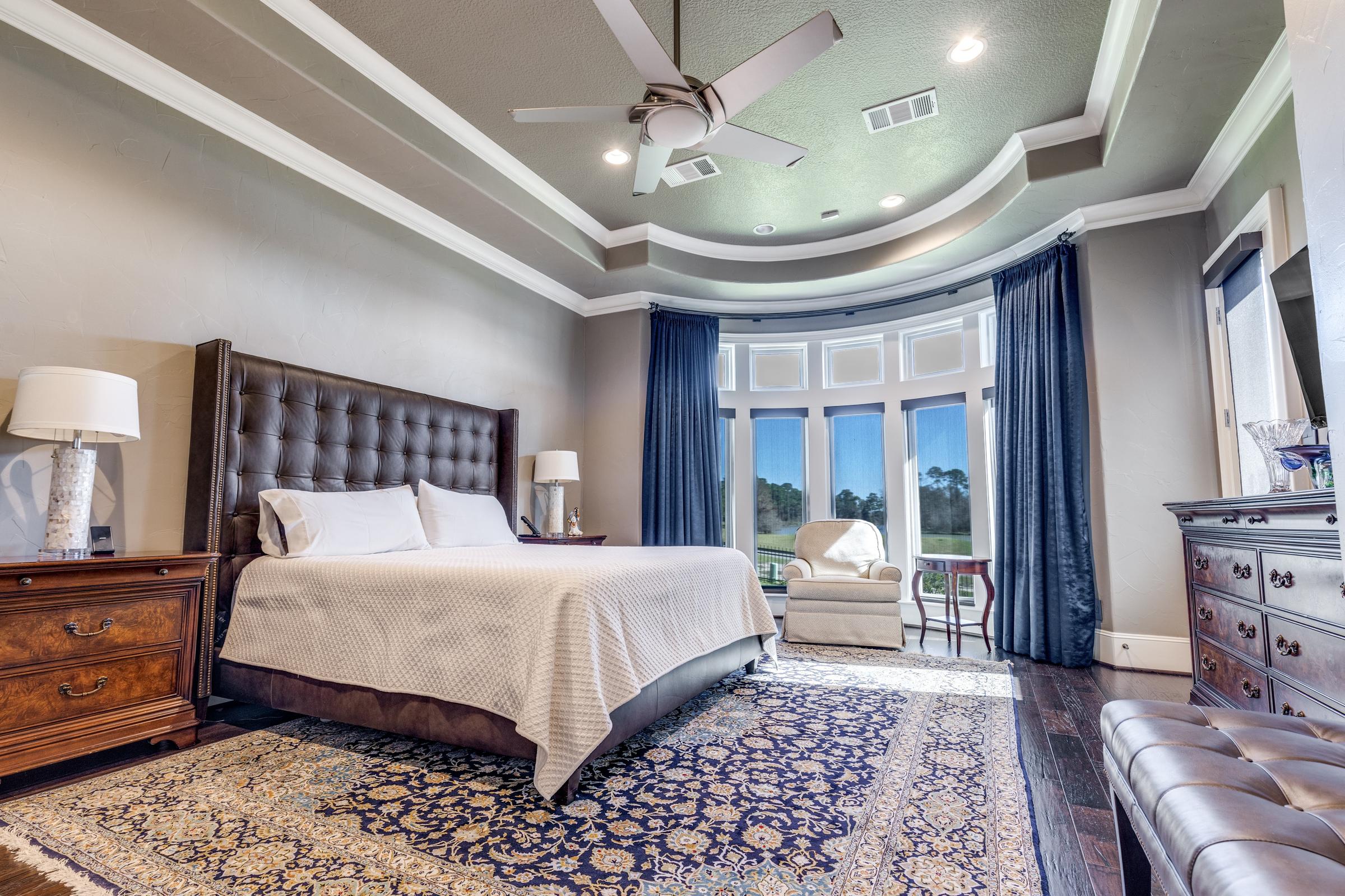 Elegant master bedroom with tufted headboard, tray ceiling, and bay windows overlooking a golf course in Humble, Texas