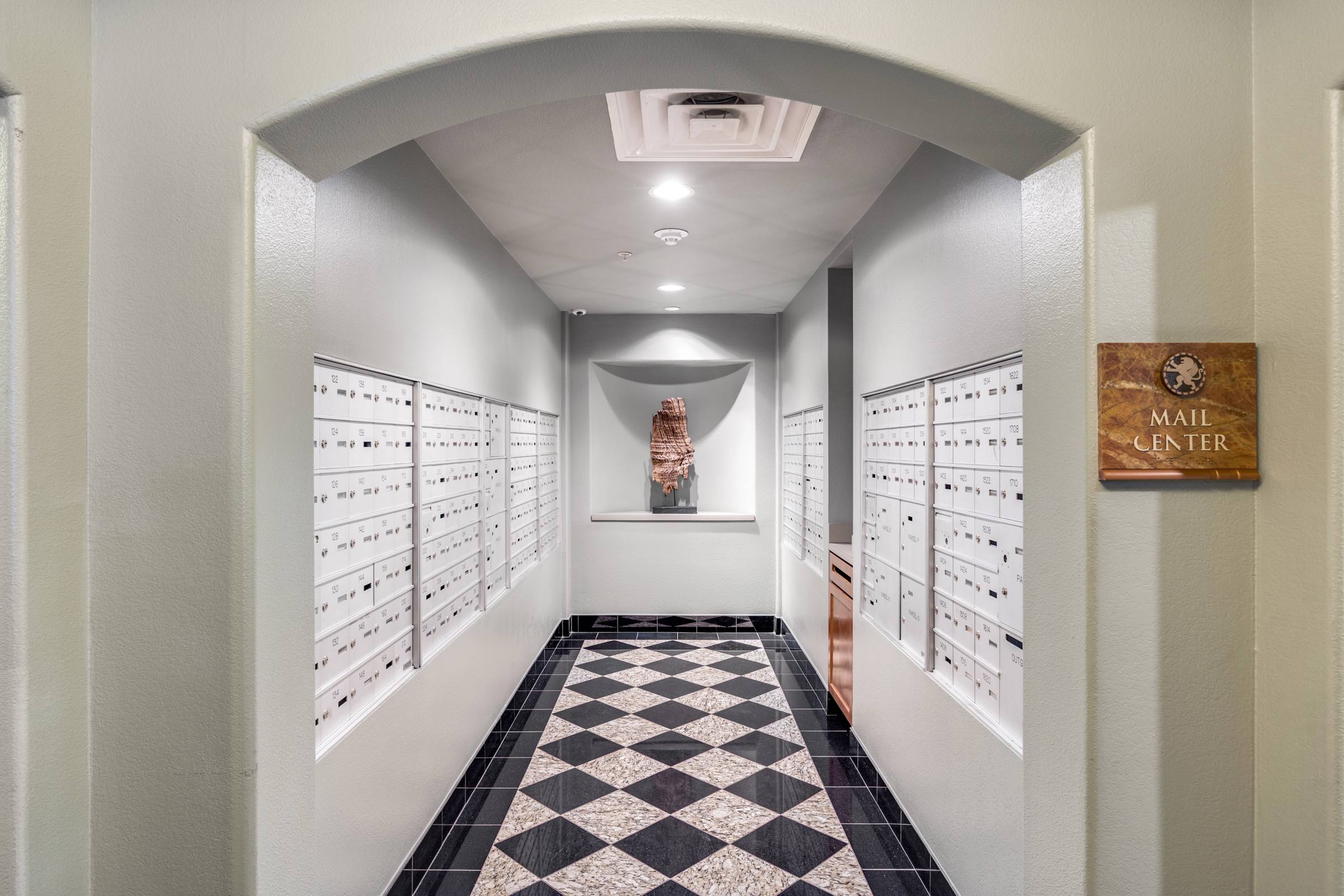 Apartment community mail center in Las Colinas, Texas, with built-in mailboxes lining an arched hallway, black-and-white checkerboard tile floor, and a Mail Center plaque on the wall.