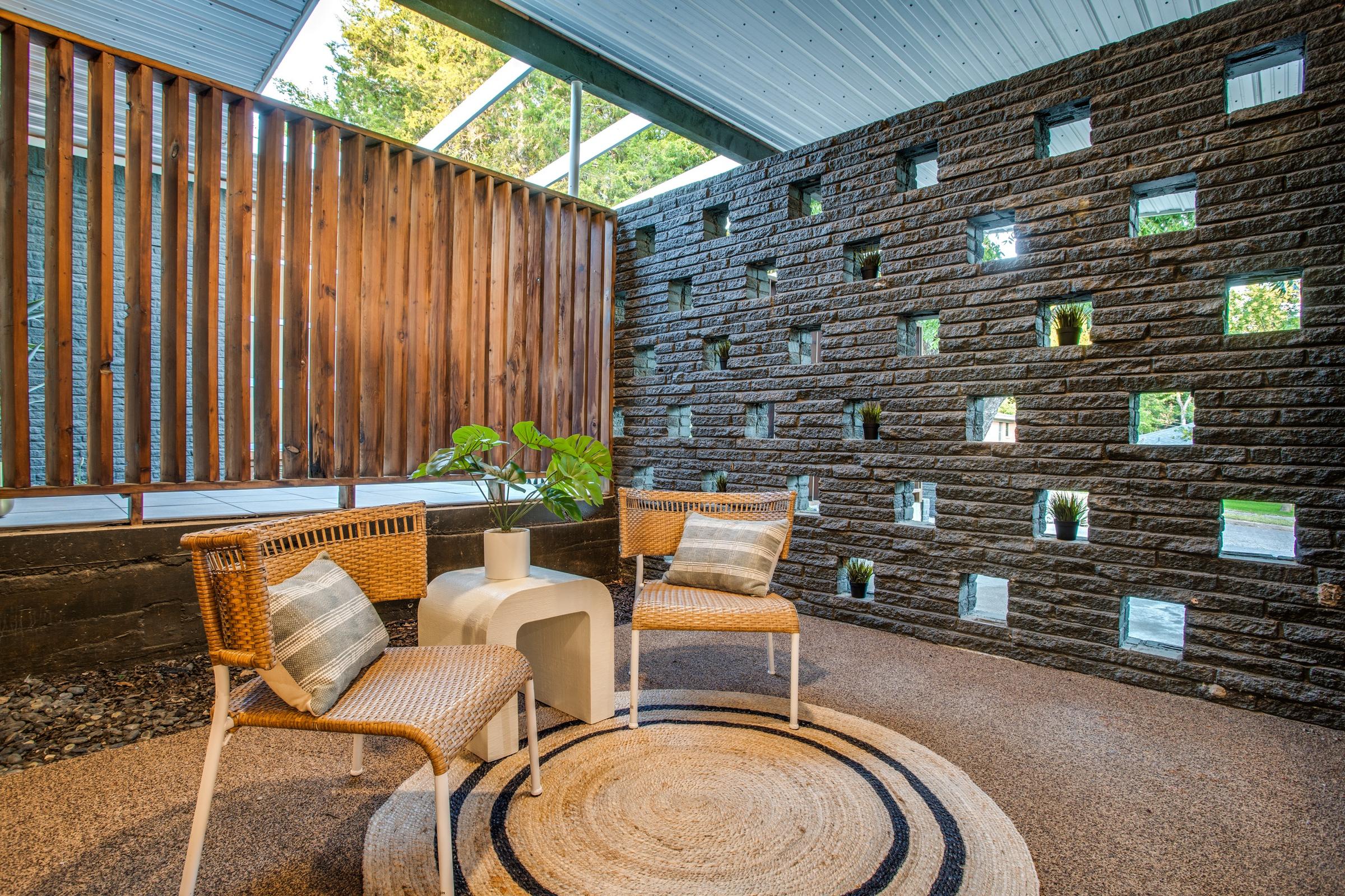 Midcentury atrium breezeway with glass block wall, natural stone, vertical wood slats, rattan chairs, and a jute area rug in a Texas estate.