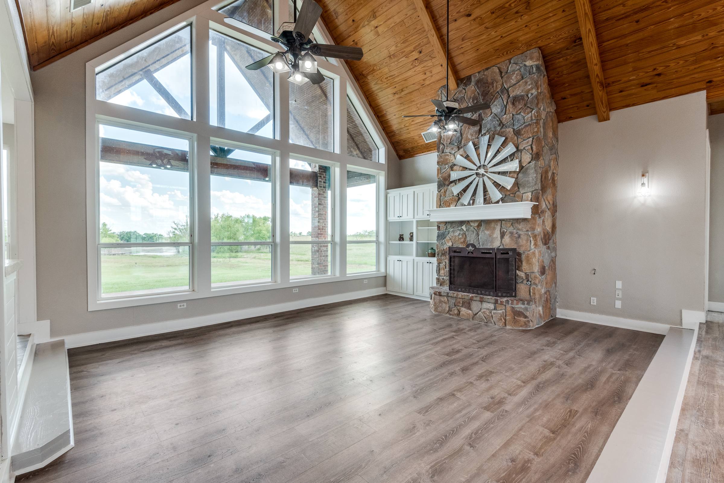 Empty living room with vaulted wood ceiling, exposed beams, stone fireplace with windmill decor, and built-in cabinets before virtual staging in Ennis, Texas