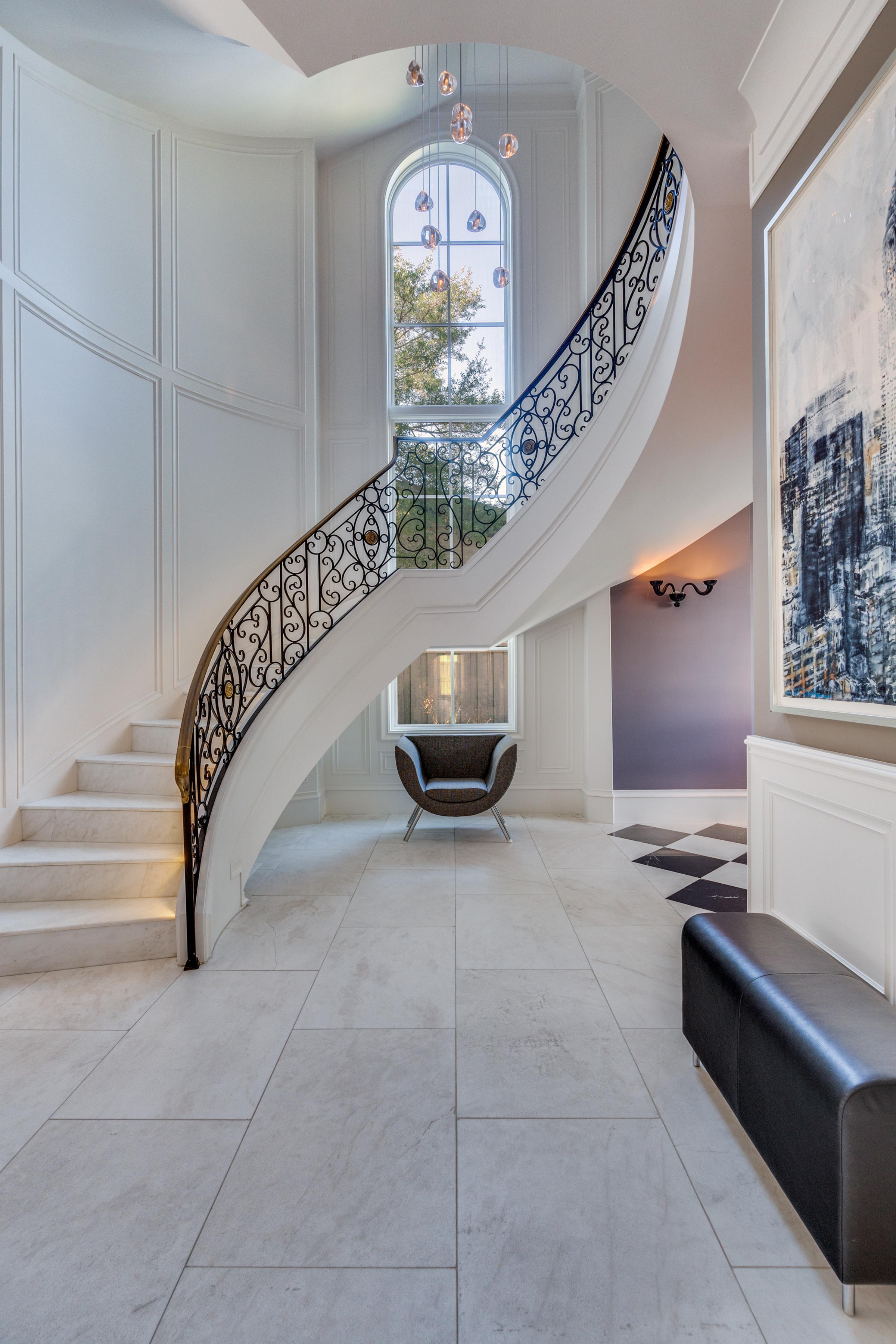 Fort Worth, TX interior portrait view of a white curved staircase with ornate wrought-iron balustrade, arched window at the landing, and contemporary chandelier above a marble tile foyer.