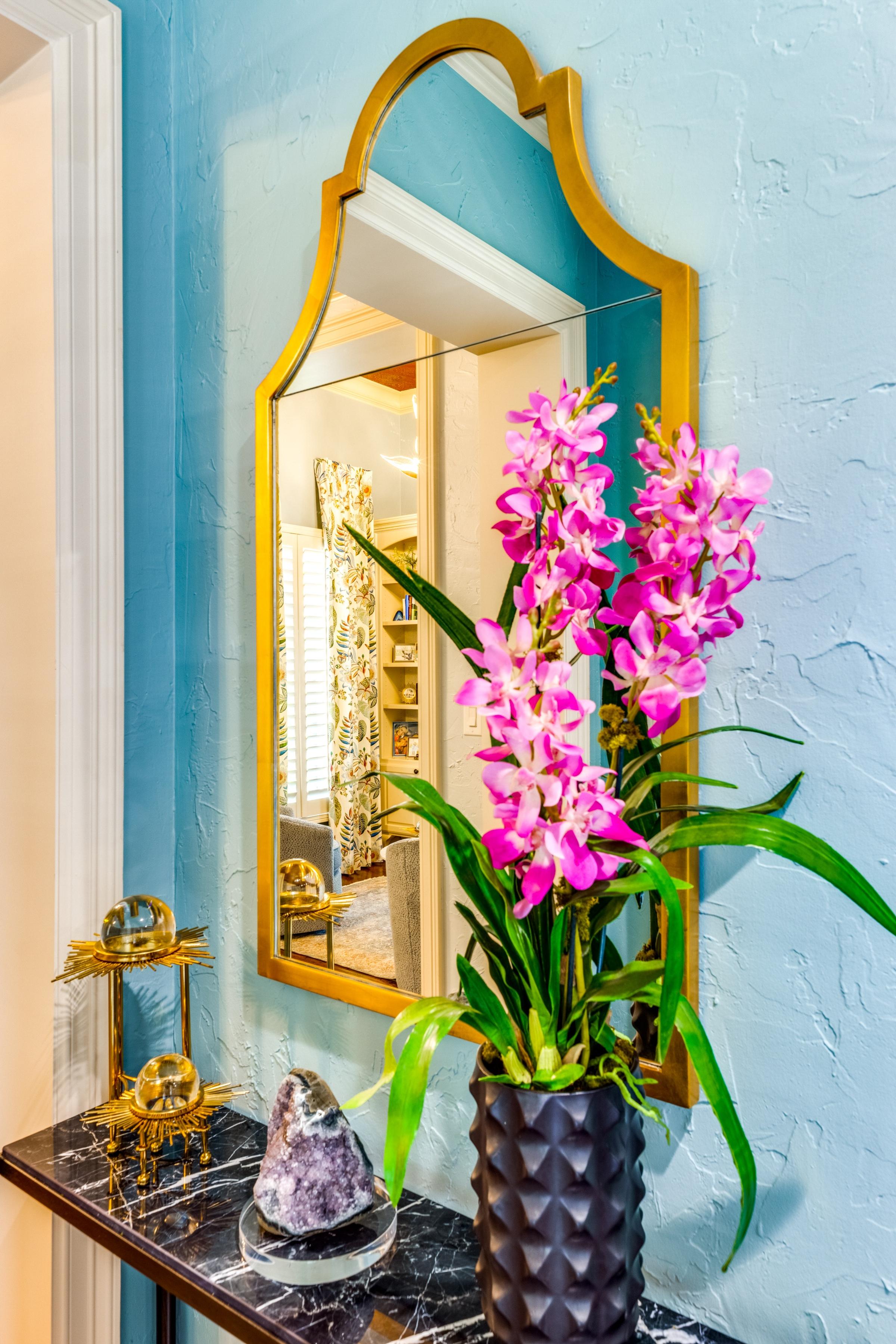 Colorful turquoise entryway with gold-framed mirror, pink orchids, amethyst crystal, and marble side table in a Texas vacation rental