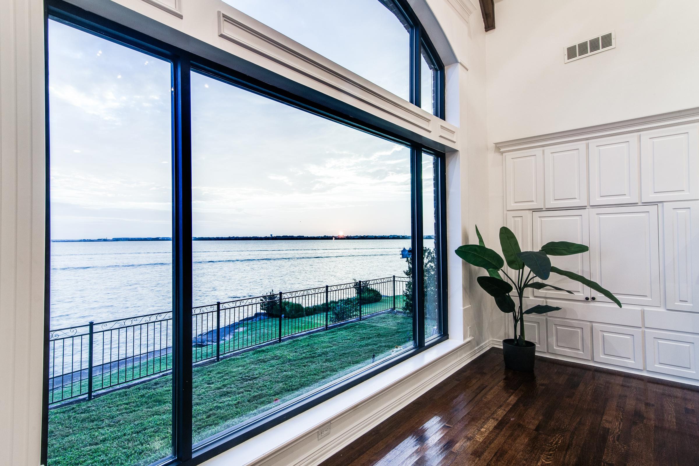 Large picture window framing an expansive lake view with dark wood floors, wainscoted walls, and a palm plant in a Texas waterfront estate.