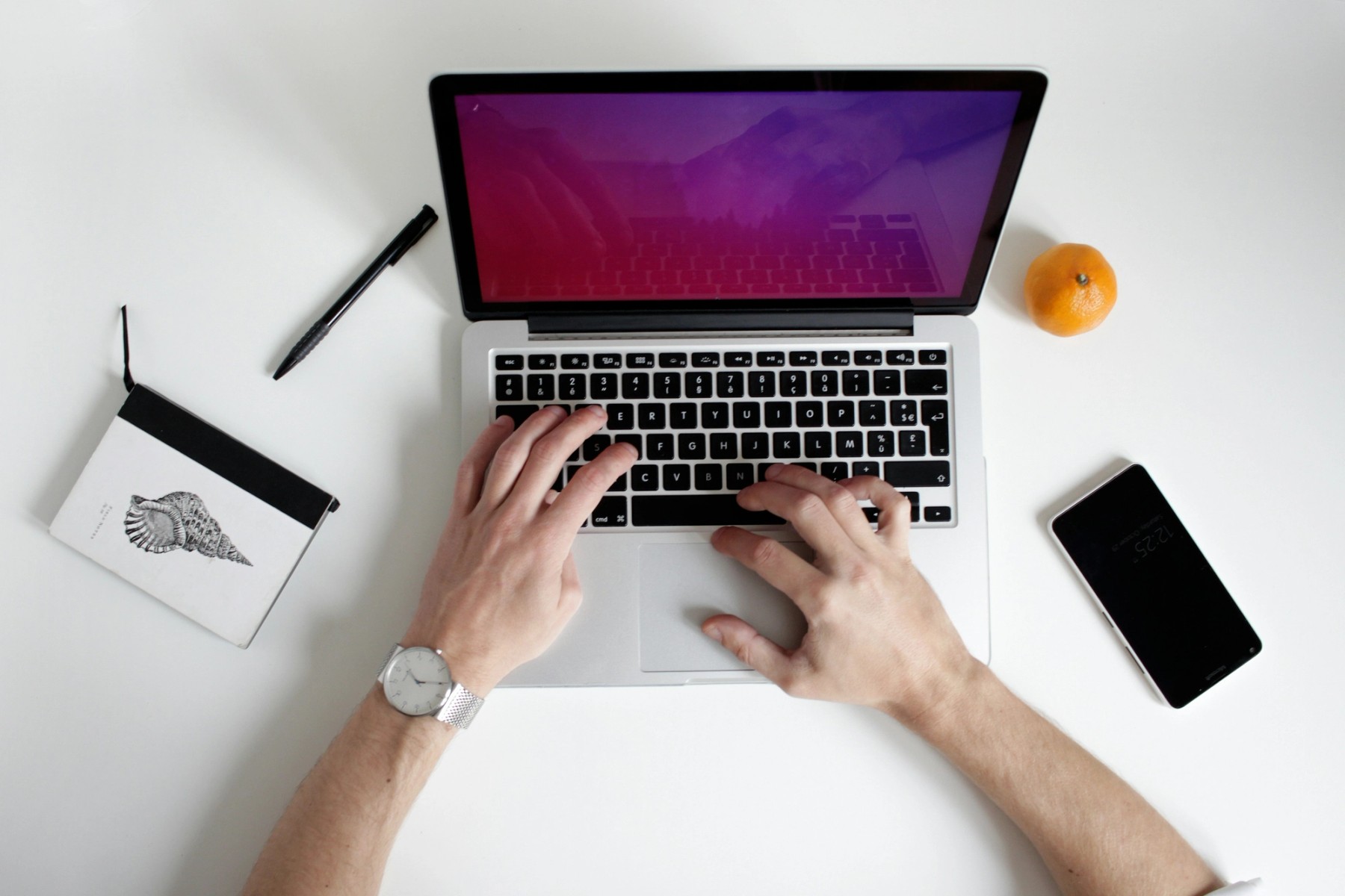 A top-down view of hands typing on a laptop keyboard, along with a sketchbook, pen, phone, and a clementine.