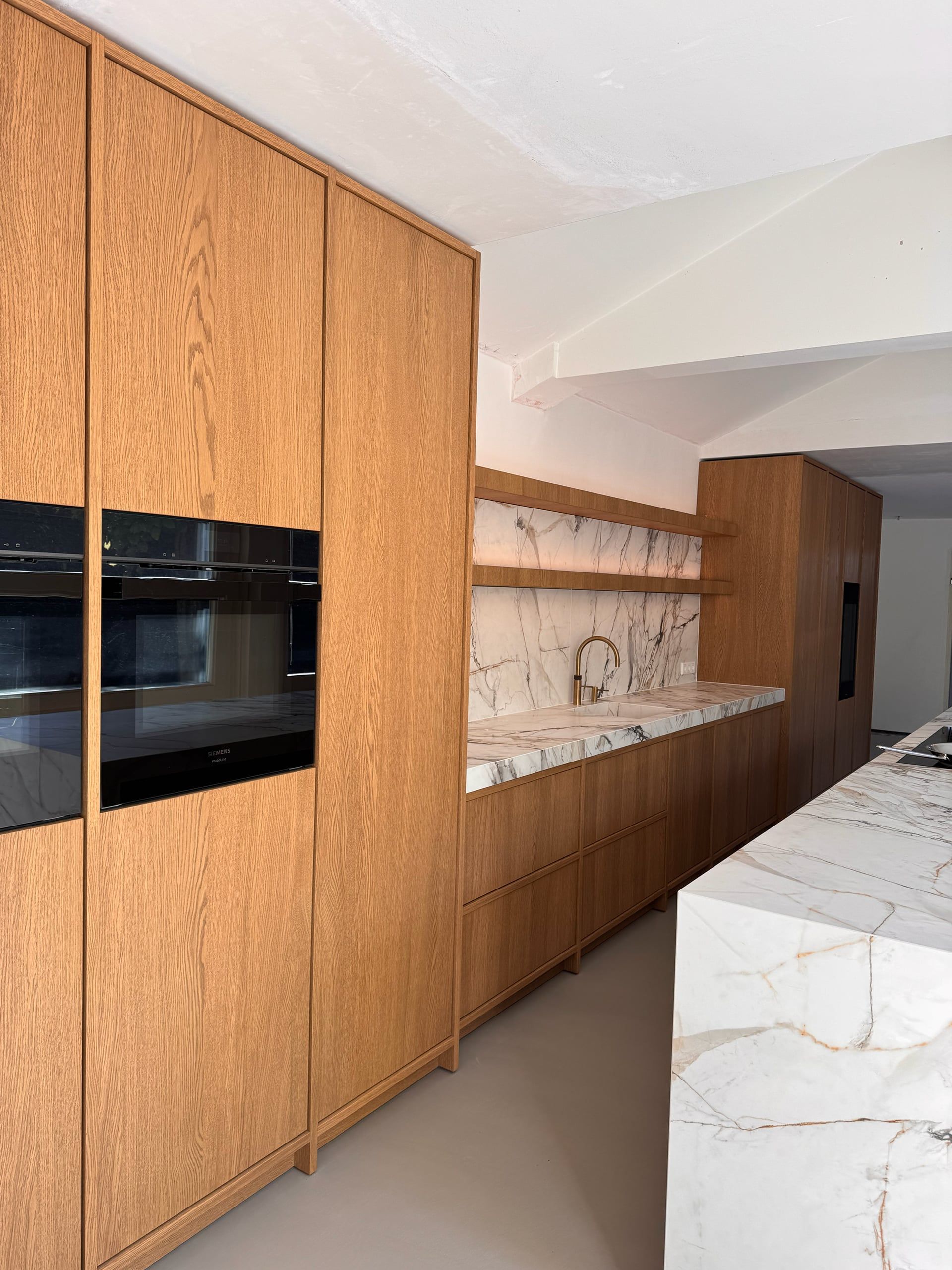Wand van hoge eiken kasten met twee ingebouwde ovens, doorlopend naar de spoelwand in marmerlook