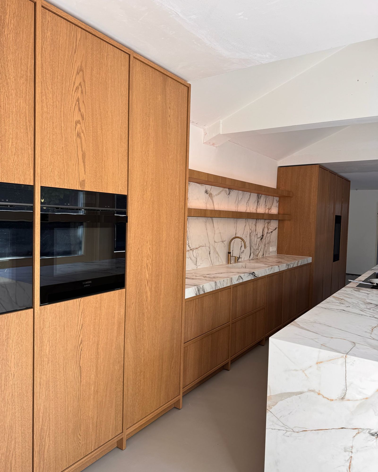 Wand van hoge eiken kasten met twee ingebouwde ovens, doorlopend naar de spoelwand in marmerlook