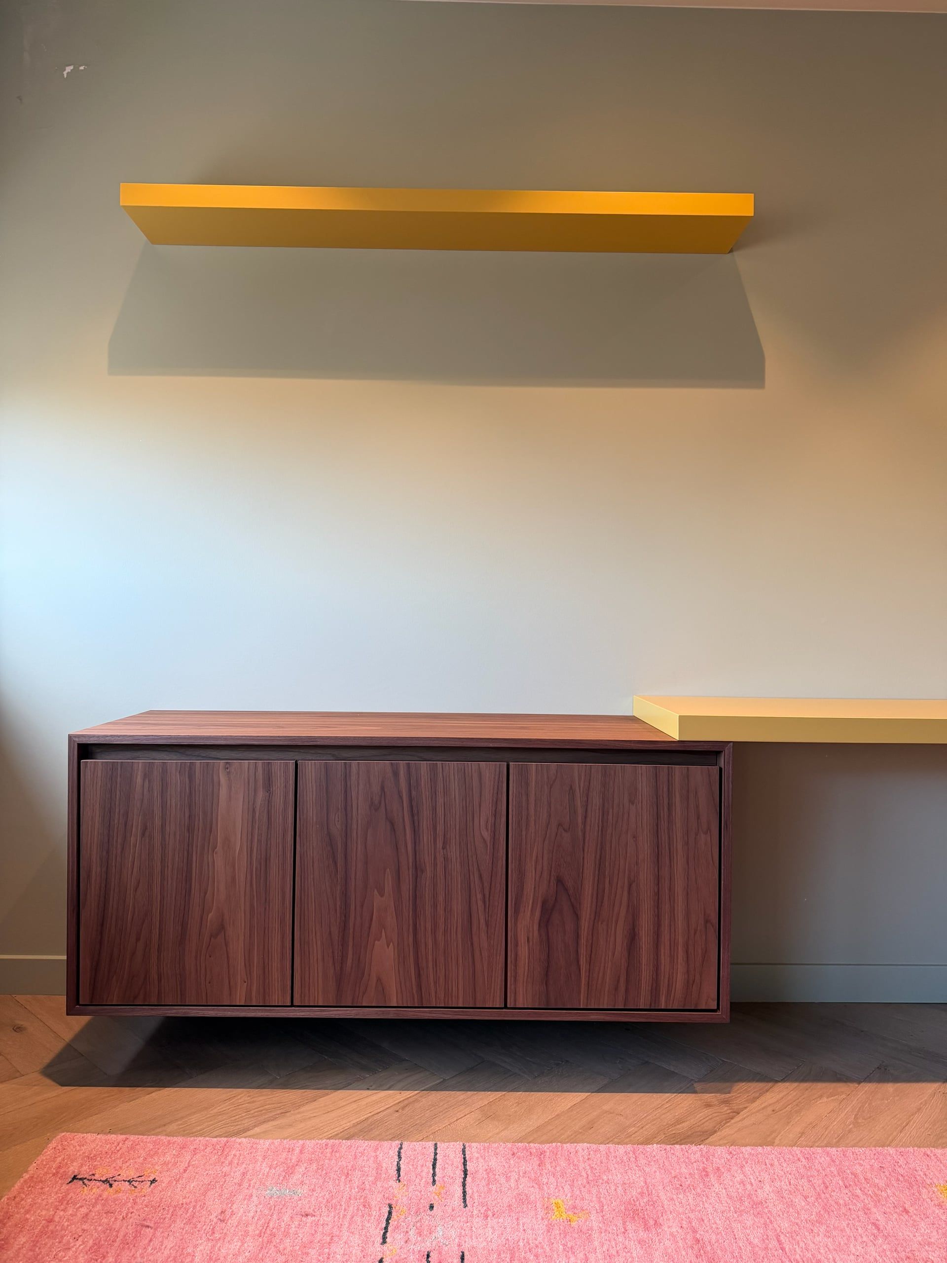 Zwevend dressoir in walnoot met drie deuren, erboven een gele wandplank en rechts het gele bureau