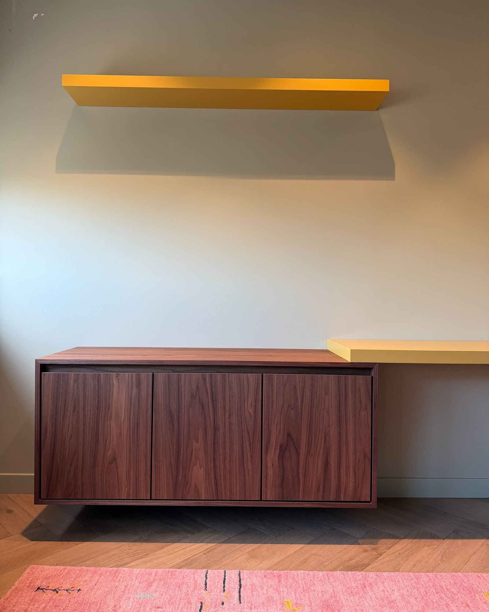 Zwevend dressoir in walnoot met drie deuren, erboven een gele wandplank en rechts het gele bureau