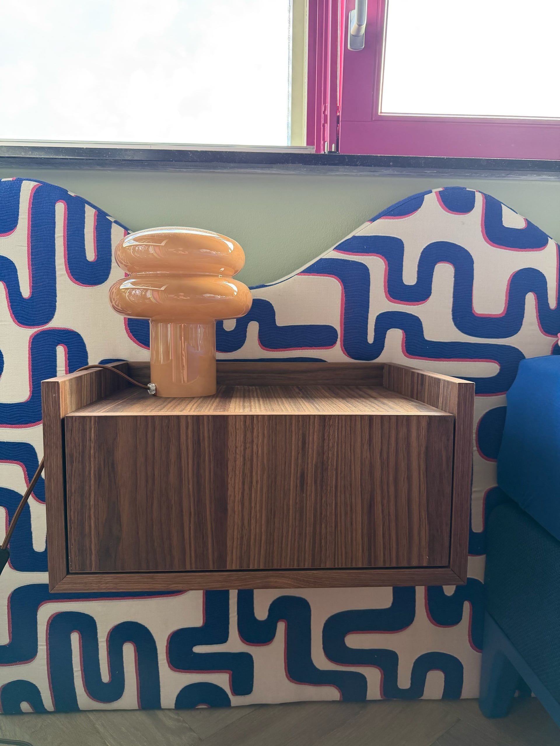 Zwevend nachtkastje in walnoot met lade en oranje lamp, tegen een golvend hoofdbord met blauw patroon