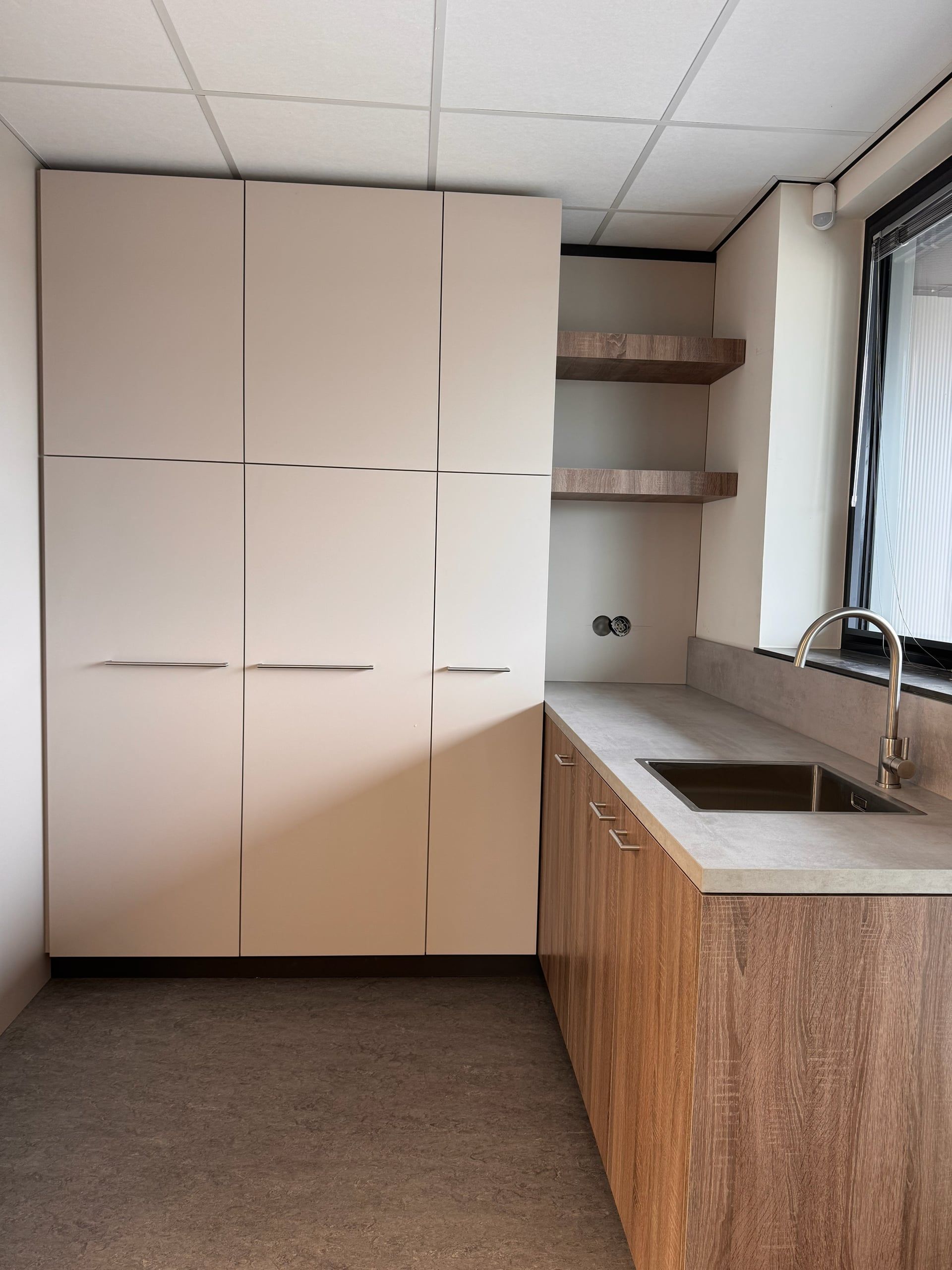 Pantrykeuken met hoge lichtgrijze kasten, twee zwevende eikenkleurige planken en onderkasten in dezelfde kleur onder een betonlook werkblad