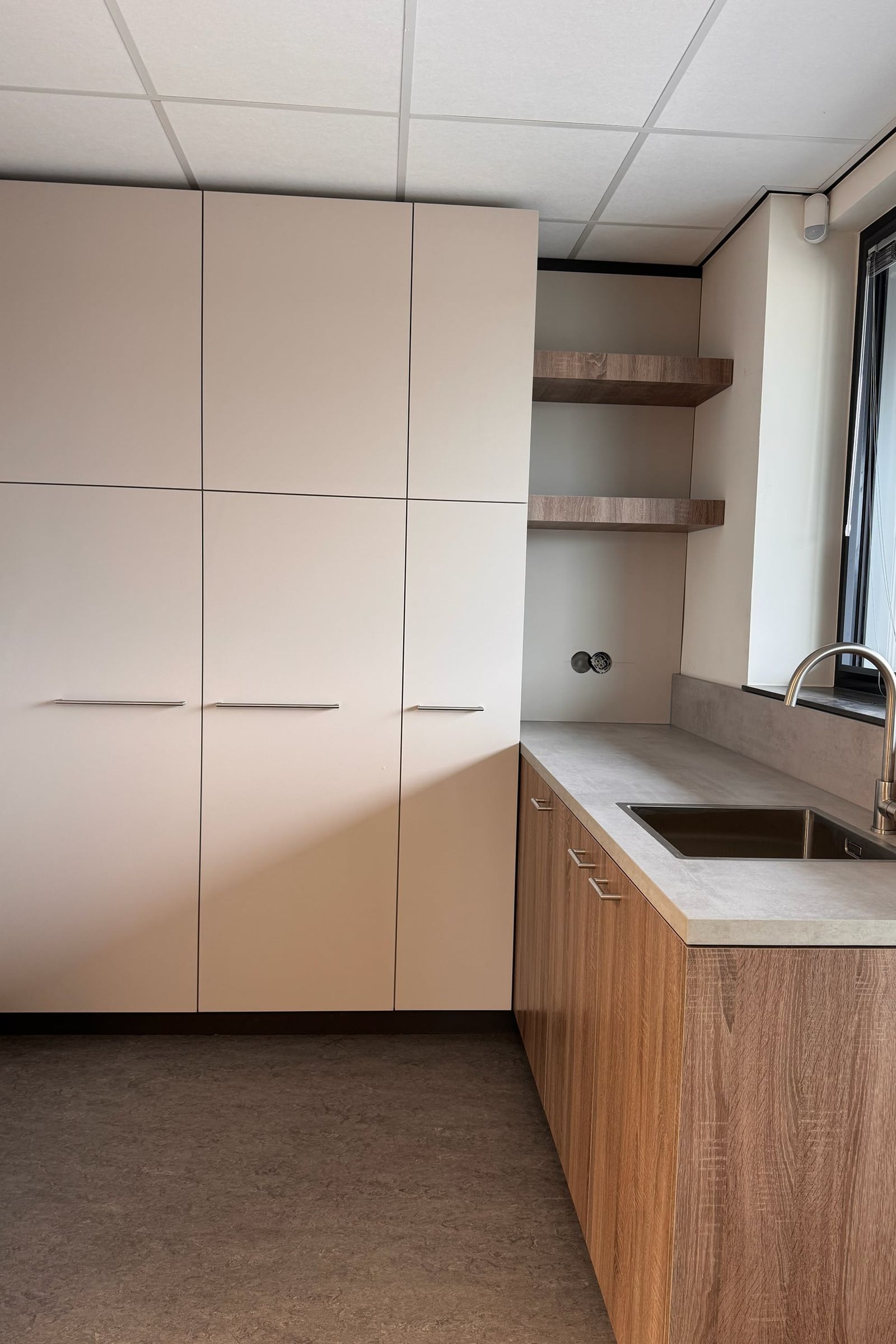 Pantrykeuken met hoge lichtgrijze kasten, twee zwevende eikenkleurige planken en onderkasten in dezelfde kleur onder een betonlook werkblad