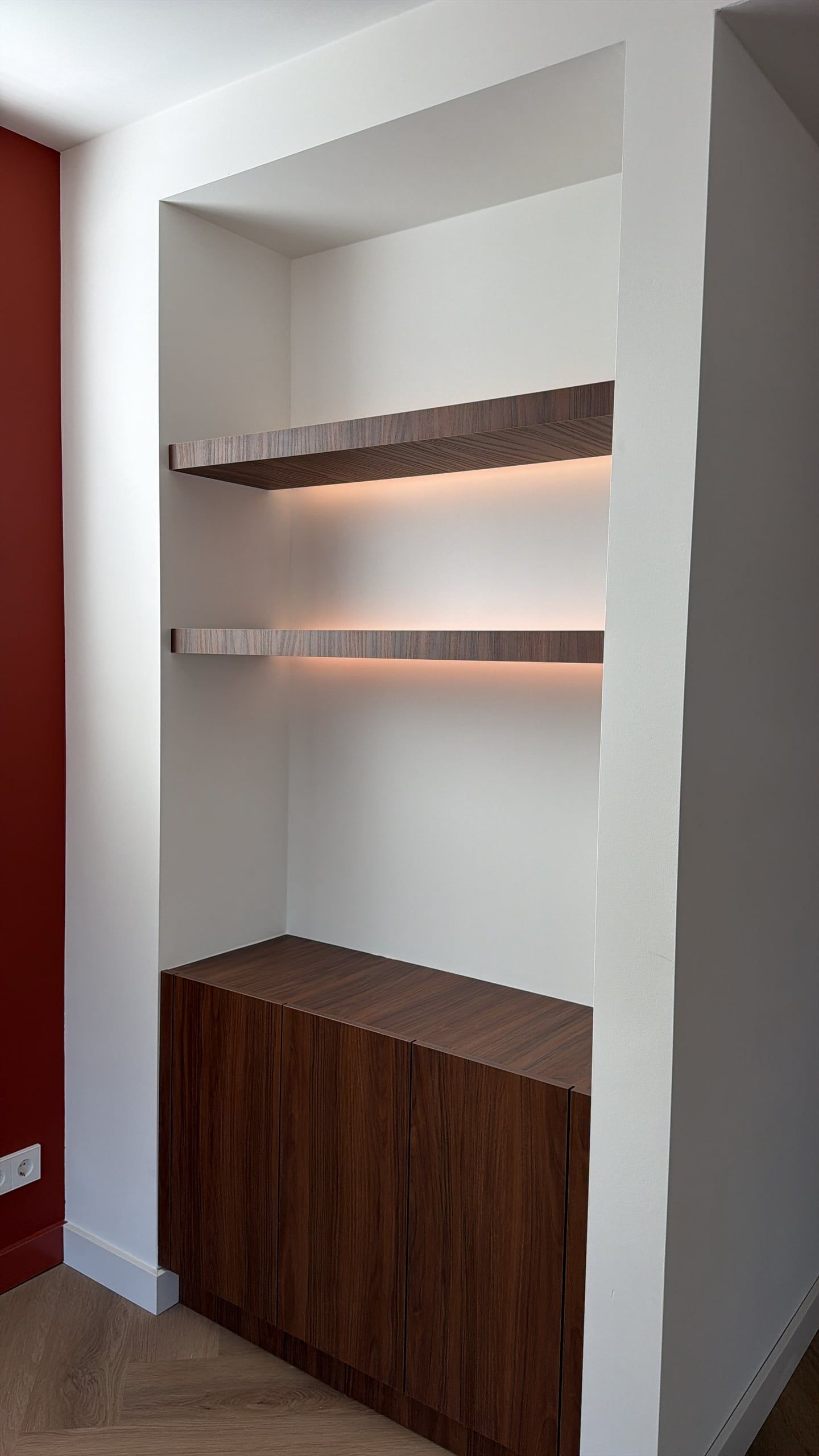 Witte wandnis met twee zwevende walnoten planken met ledverlichting boven een lage walnoten kast, naast een donkerrode wand