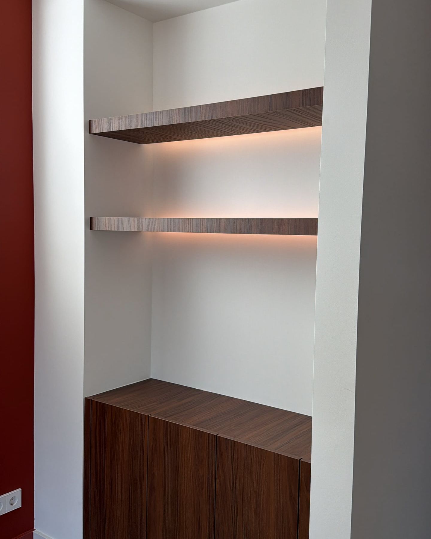 Witte wandnis met twee zwevende walnoten planken met ledverlichting boven een lage walnoten kast, naast een donkerrode wand