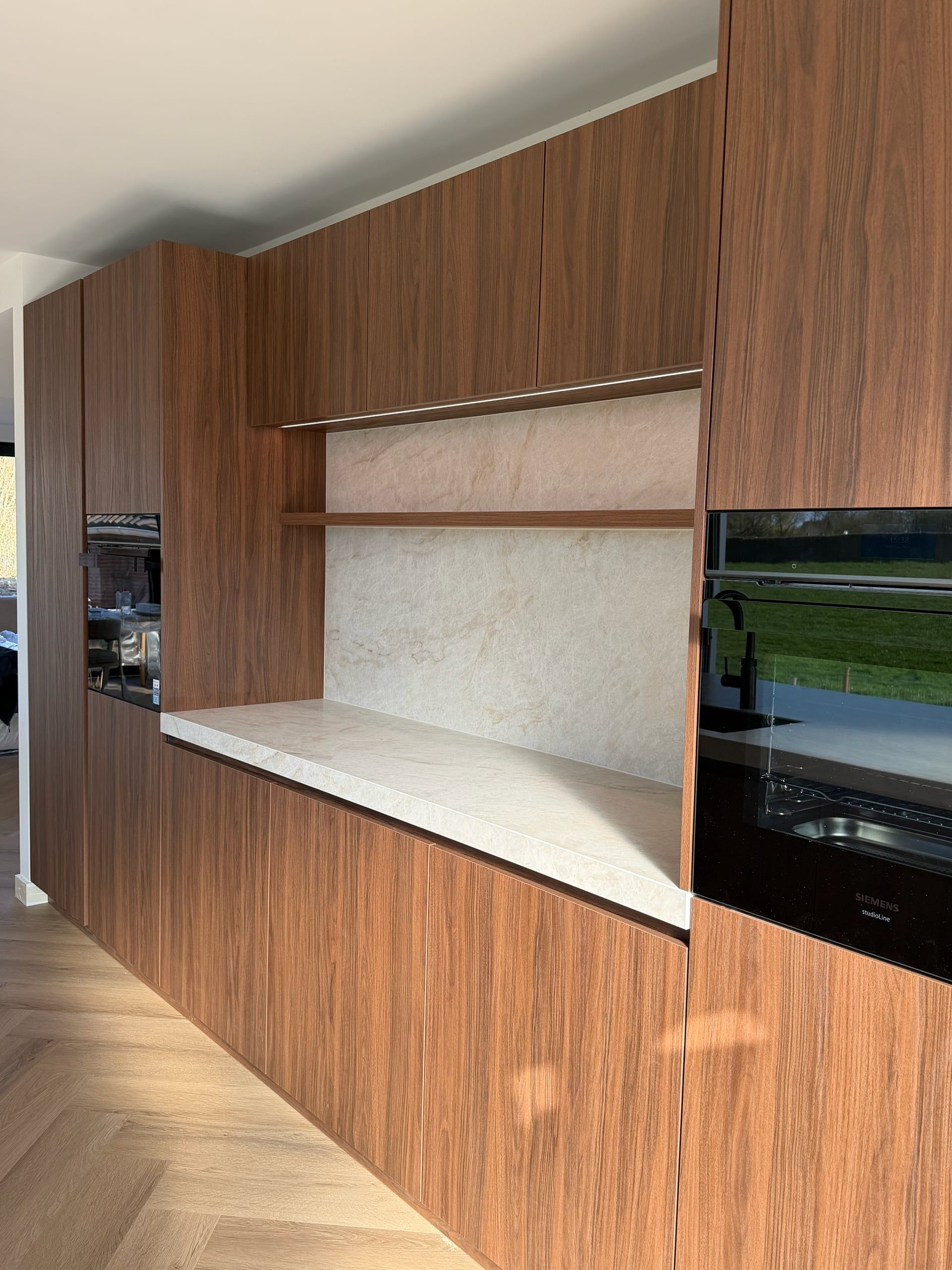 Keukenwand in walnoot met ovens en een nis met stenen achterwand, walnoten plank en ledverlichting boven het werkblad