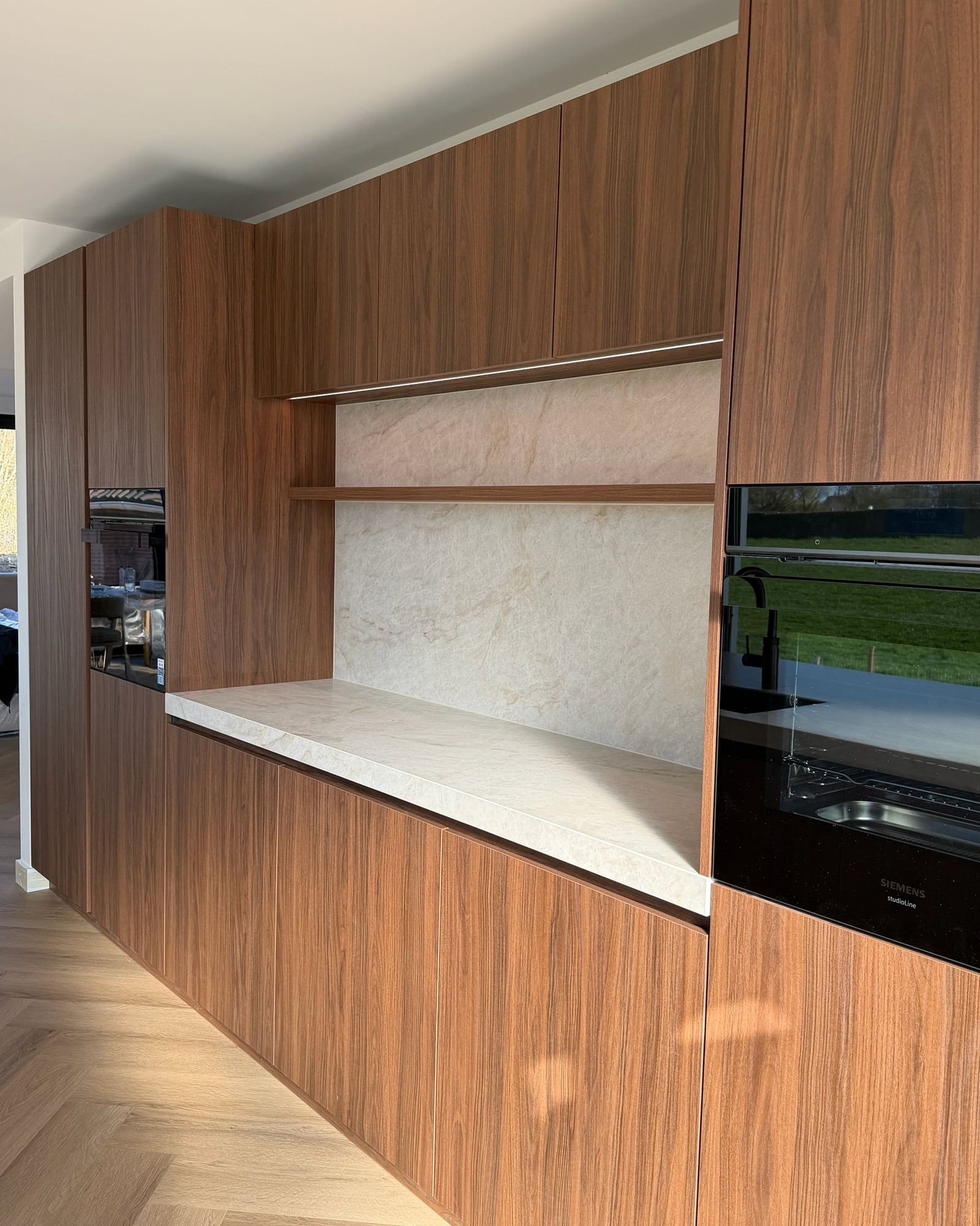 Keukenwand in walnoot met ovens en een nis met stenen achterwand, walnoten plank en ledverlichting boven het werkblad