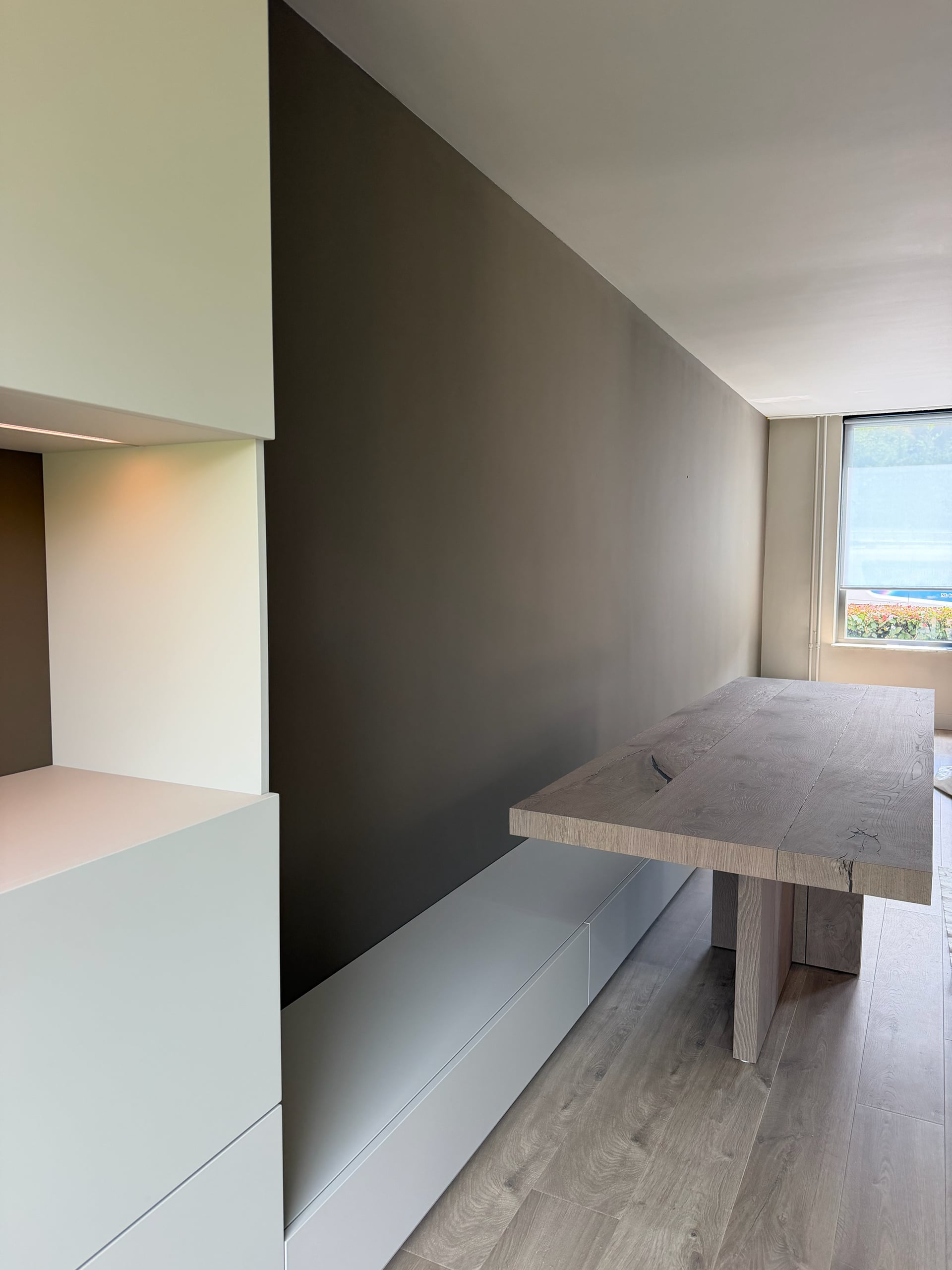 Lichte kastenwand met verlichte nis, lage witte bank en massief eiken tafel langs een taupe wand