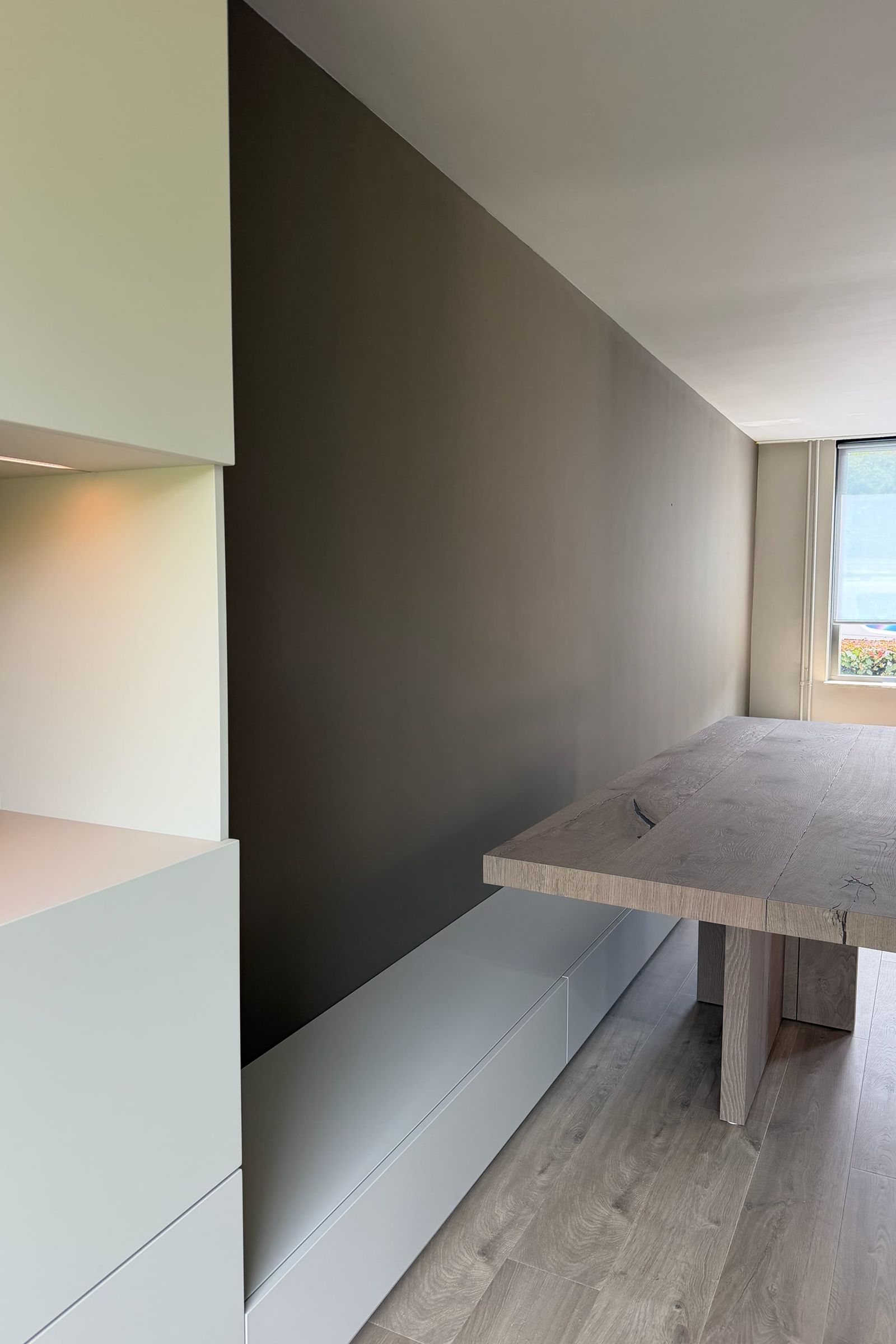 Lichte kastenwand met verlichte nis, lage witte bank en massief eiken tafel langs een taupe wand