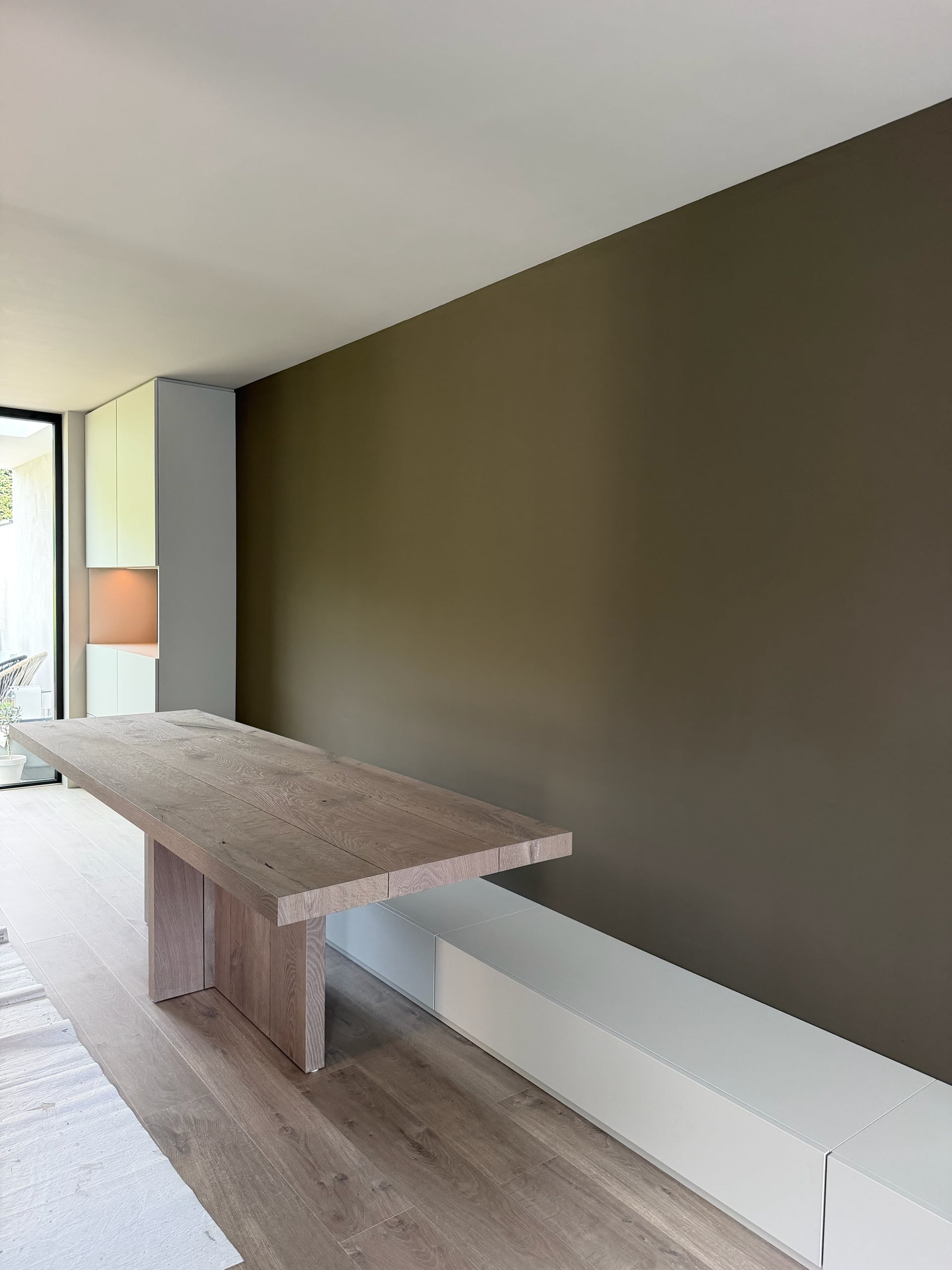 Massief eiken eettafel en lage witte bank met laden tegen een taupe wand, achterin de kastenwand