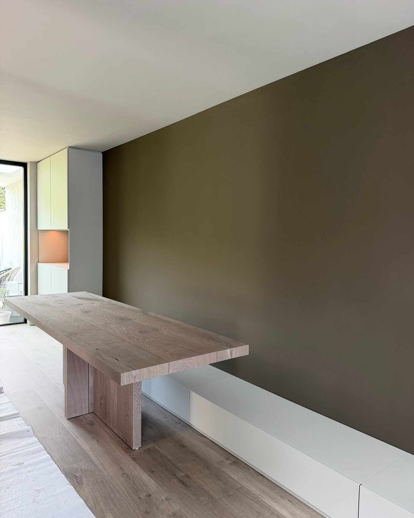 Massief eiken eettafel en lage witte bank met laden tegen een taupe wand, achterin de kastenwand