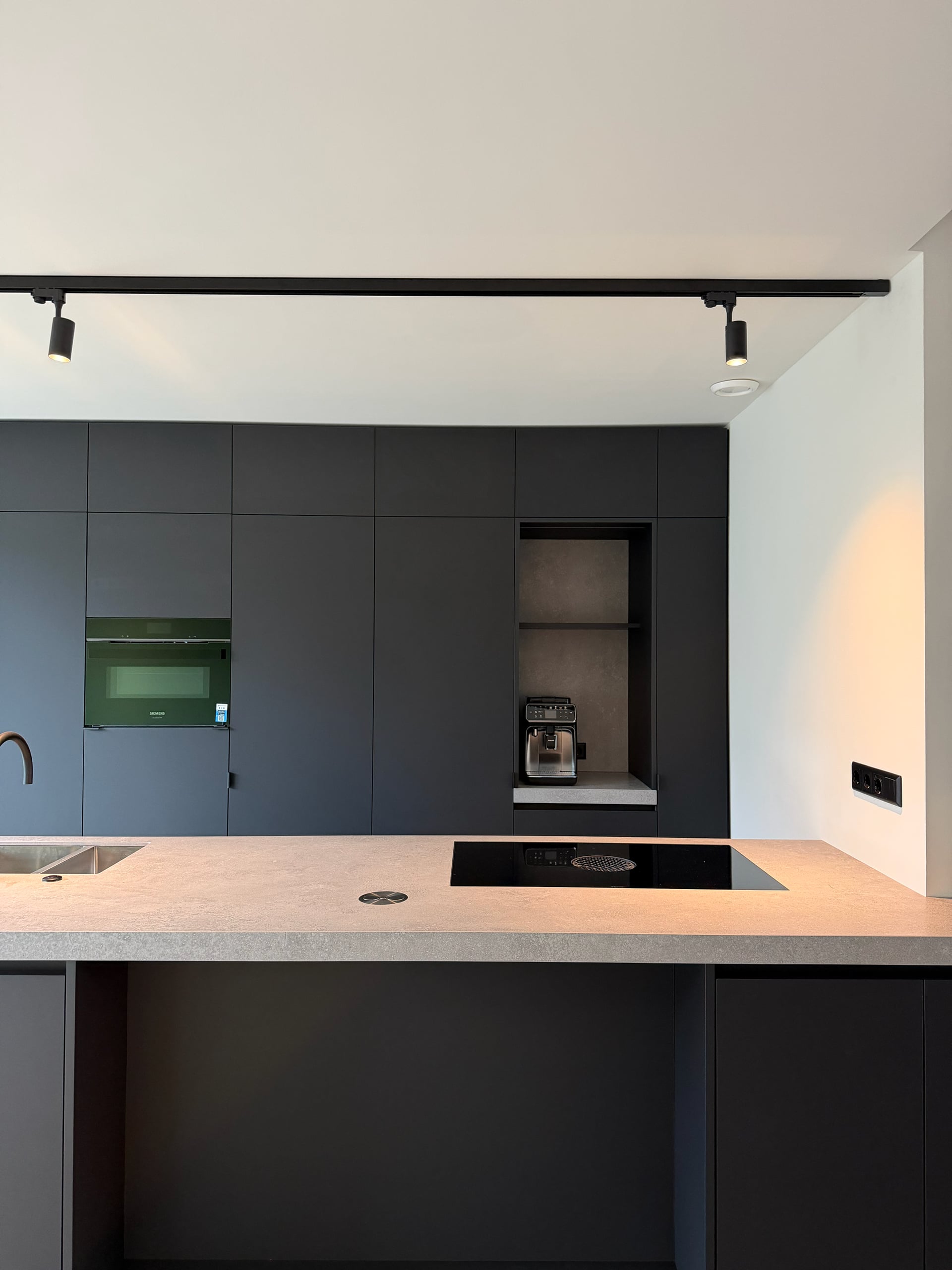 Mat zwarte kastenwand tot het plafond met oven en open koffienis, kookeiland met inductiekookplaat op de voorgrond