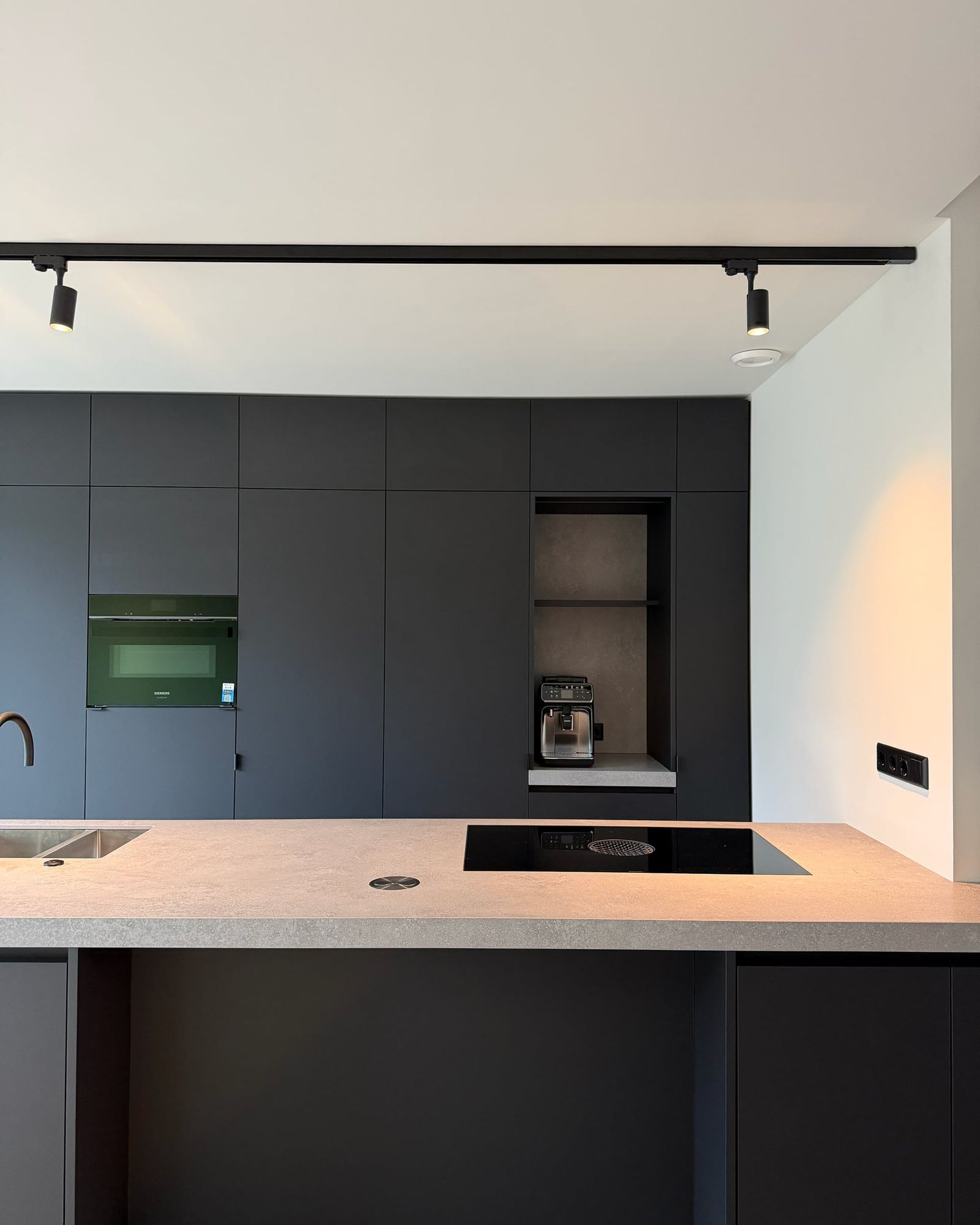 Mat zwarte kastenwand tot het plafond met oven en open koffienis, kookeiland met inductiekookplaat op de voorgrond