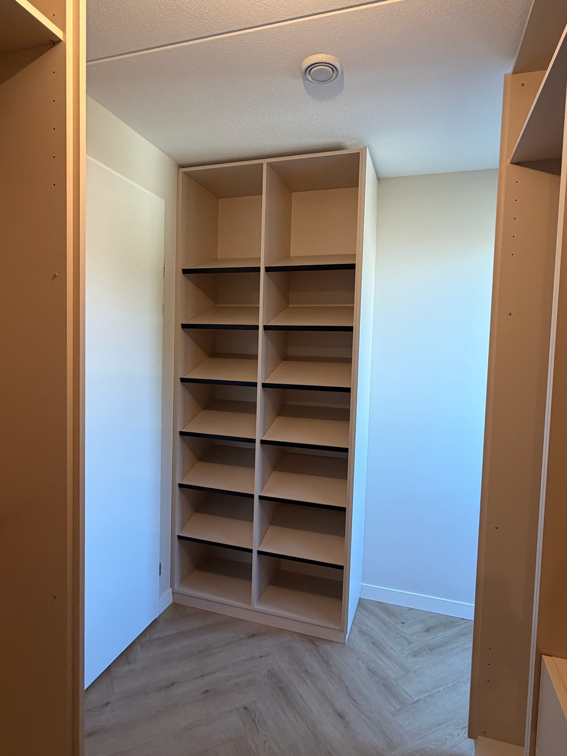 Hoge open schoenenkast in licht beige met schuine legplanken met zwarte rand, aan het eind van de inloopkast