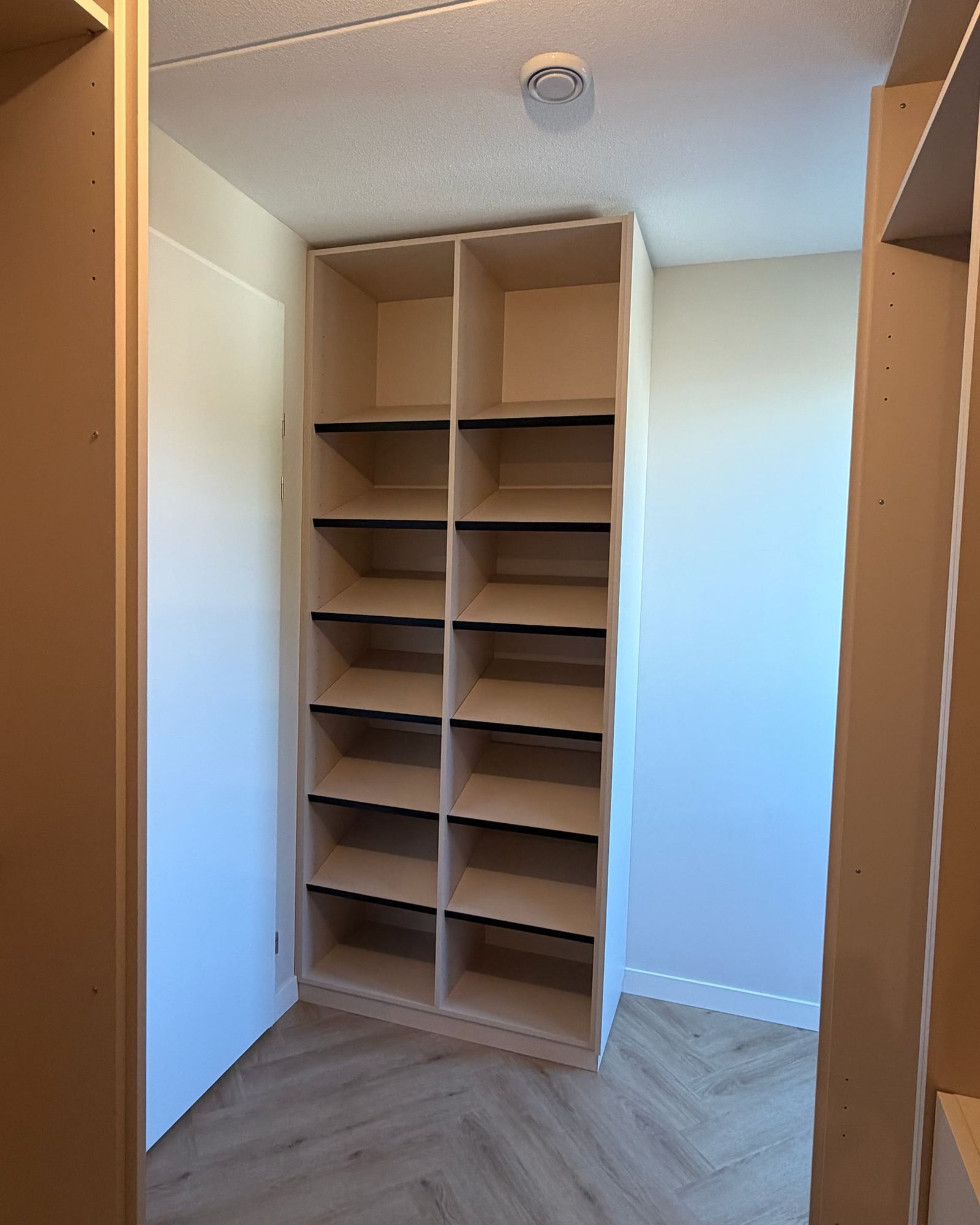 Hoge open schoenenkast in licht beige met schuine legplanken met zwarte rand, aan het eind van de inloopkast