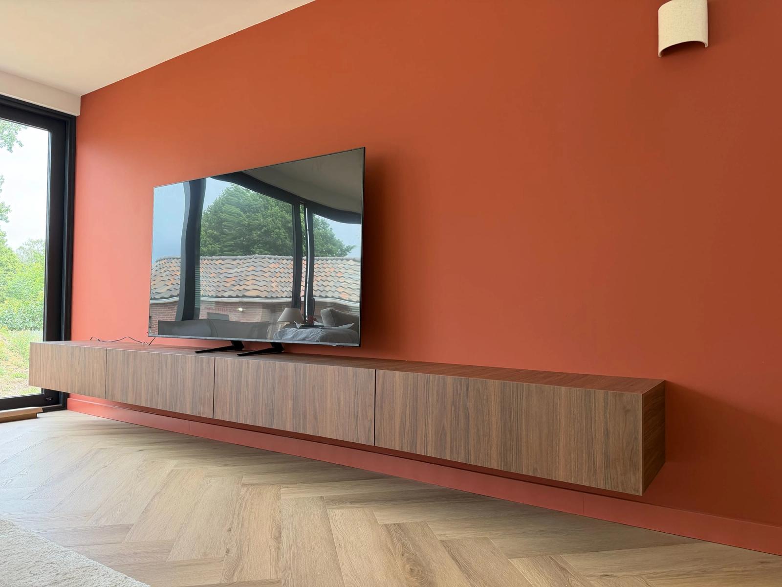 Lang zwevend tv-meubel in walnoot met greeploze fronten tegen een terracotta wand, boven een visgraatvloer