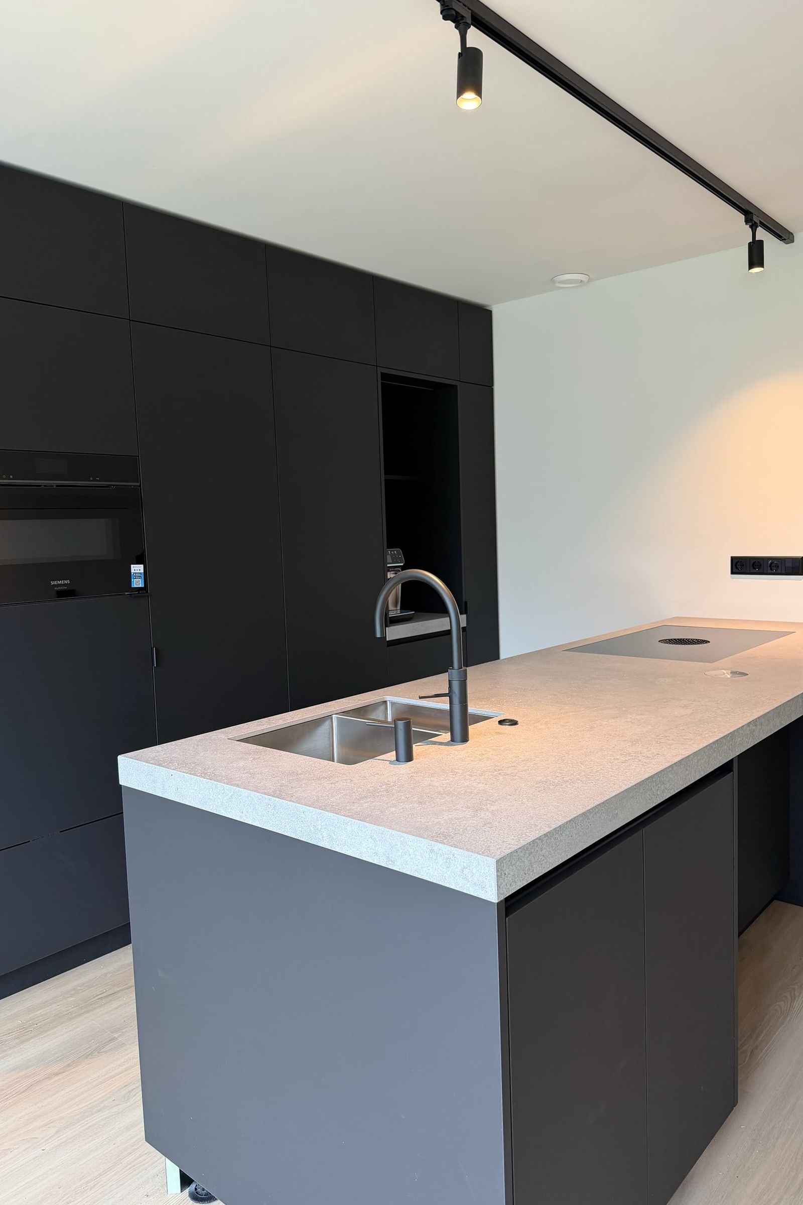 Kookeiland met dik grijs werkblad in steenlook en spoelbak voor een mat zwarte kastenwand met oven