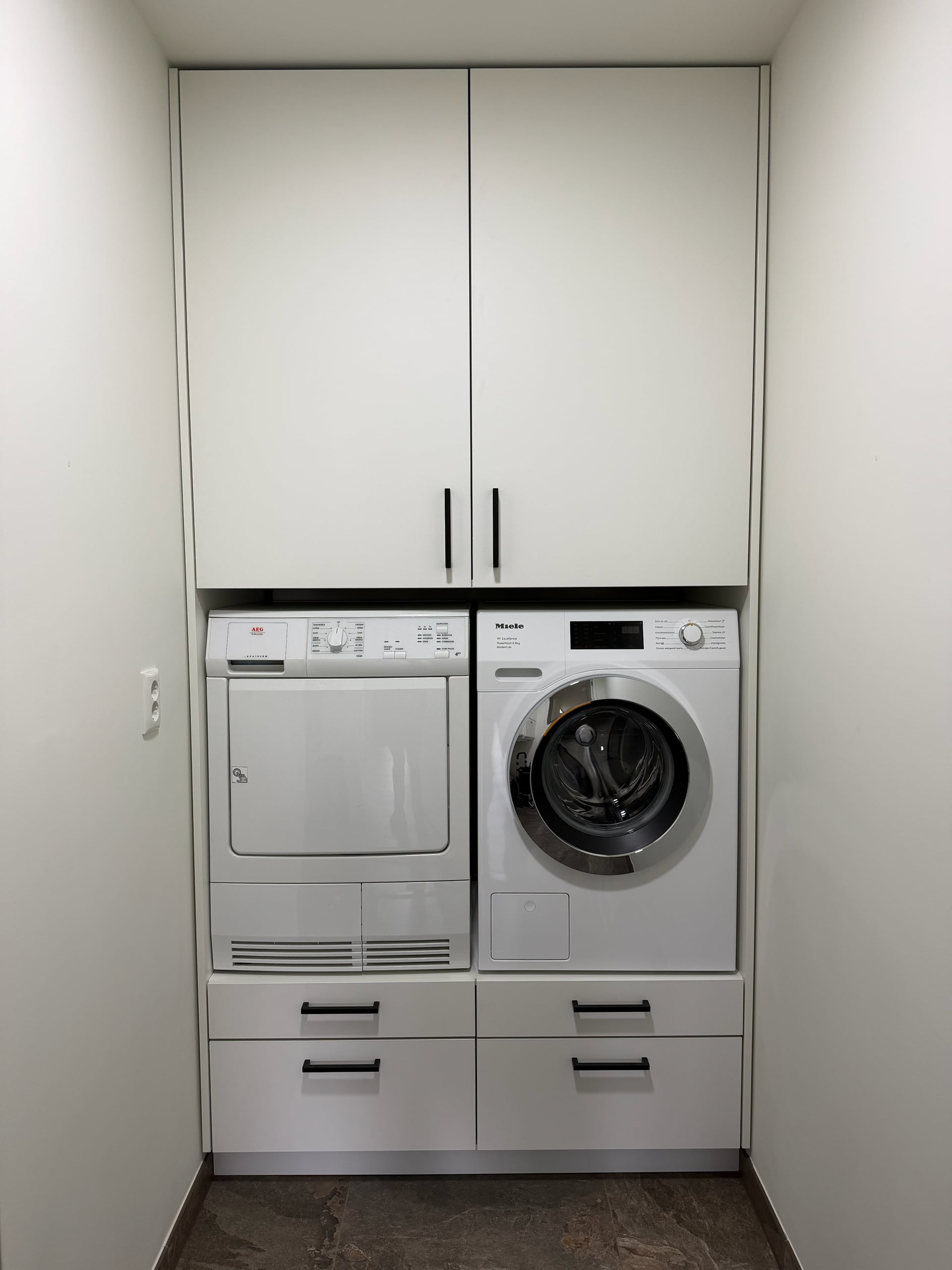 Witte inbouwkast met wasmachine en droger naast elkaar op een verhoging met lades, bovenkasten met zwarte grepen
