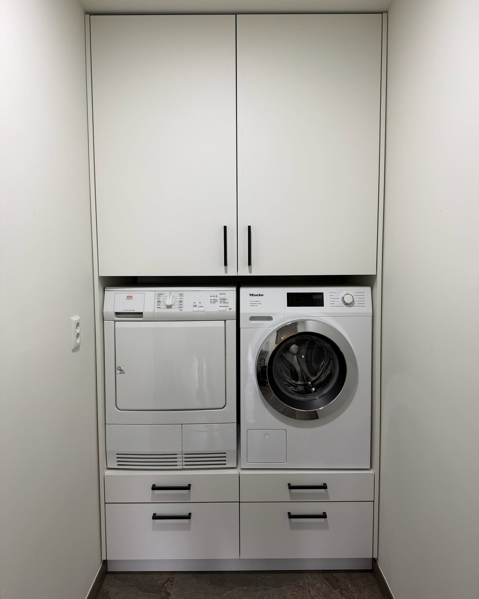 Witte inbouwkast met wasmachine en droger naast elkaar op een verhoging met lades, bovenkasten met zwarte grepen