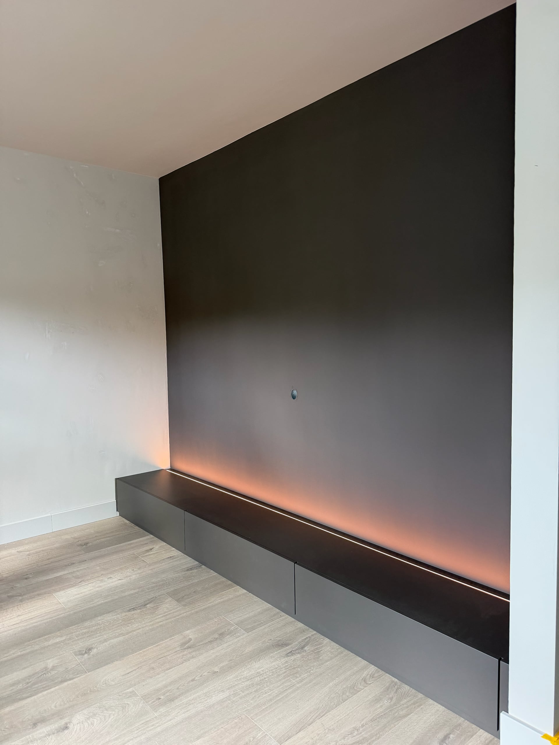 Laag antracietkleurig tv-meubel met laden over de volle breedte van een donkere wand, met ledstrip erachter