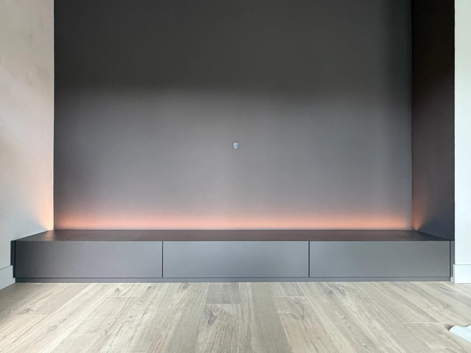 Laag antracietkleurig tv-meubel met drie laden en indirecte ledverlichting tegen een donkere wand