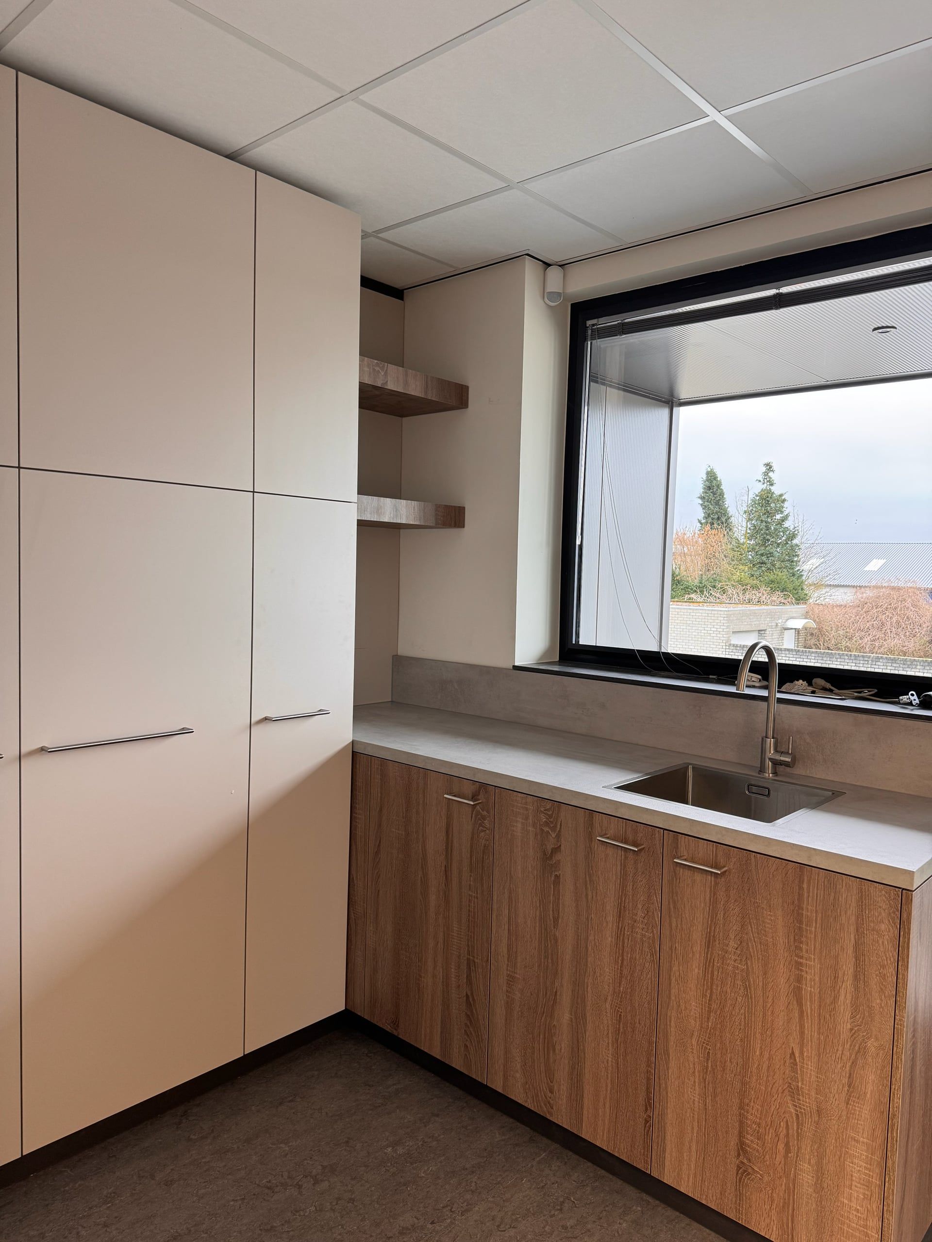 Keukenblok met eikenkleurige onderkasten, betonlook werkblad en rvs spoelbak onder een groot raam, links hoge lichtgrijze kasten