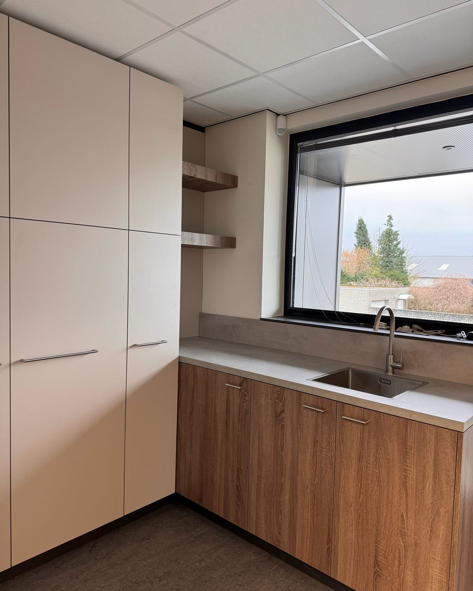 Keukenblok met eikenkleurige onderkasten, betonlook werkblad en rvs spoelbak onder een groot raam, links hoge lichtgrijze kasten