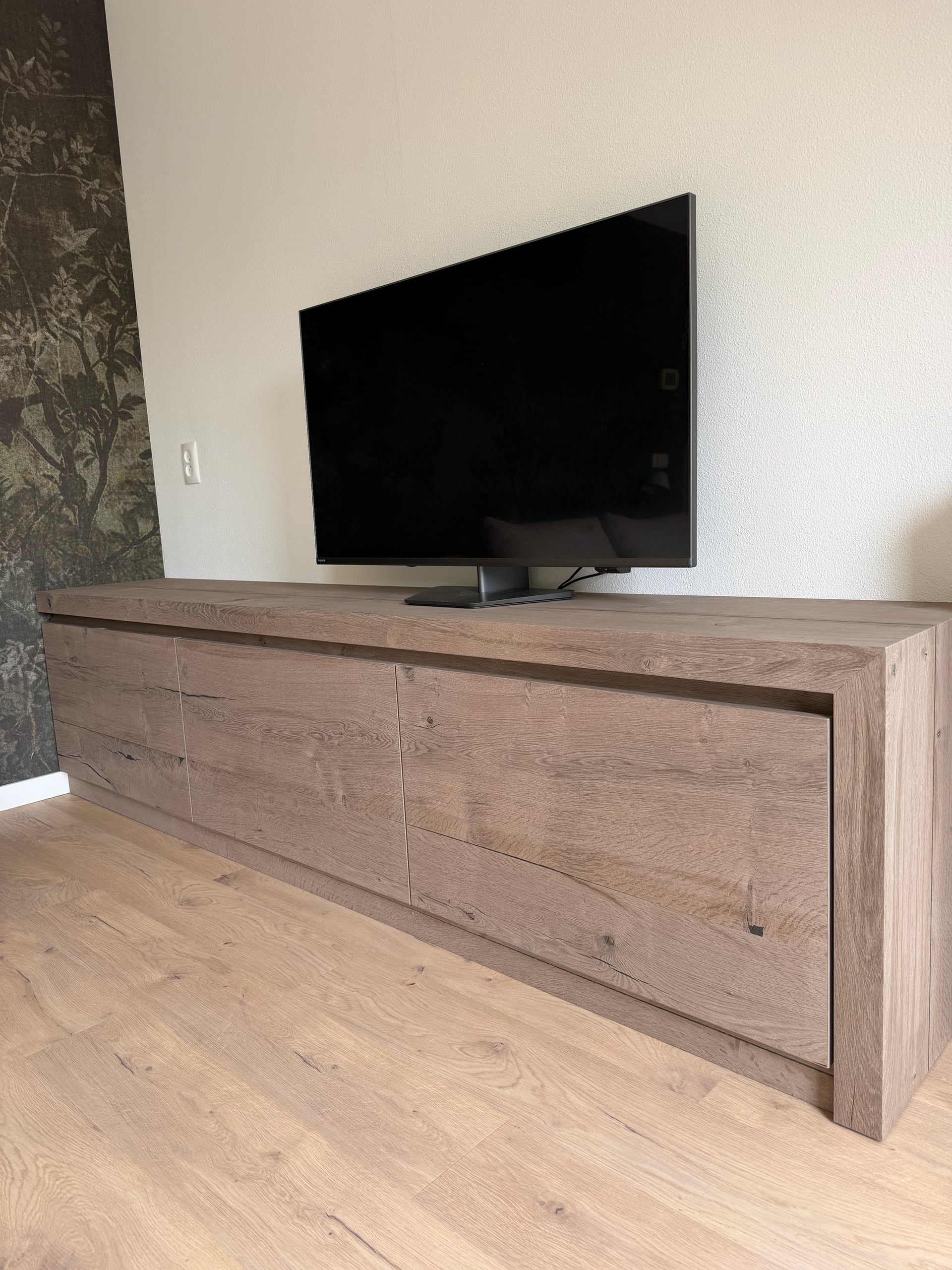Laag tv-dressoir in eiken met noesten en scheurtjes, greeploze fronten en doorlopende zijkant, tegen een witte wand