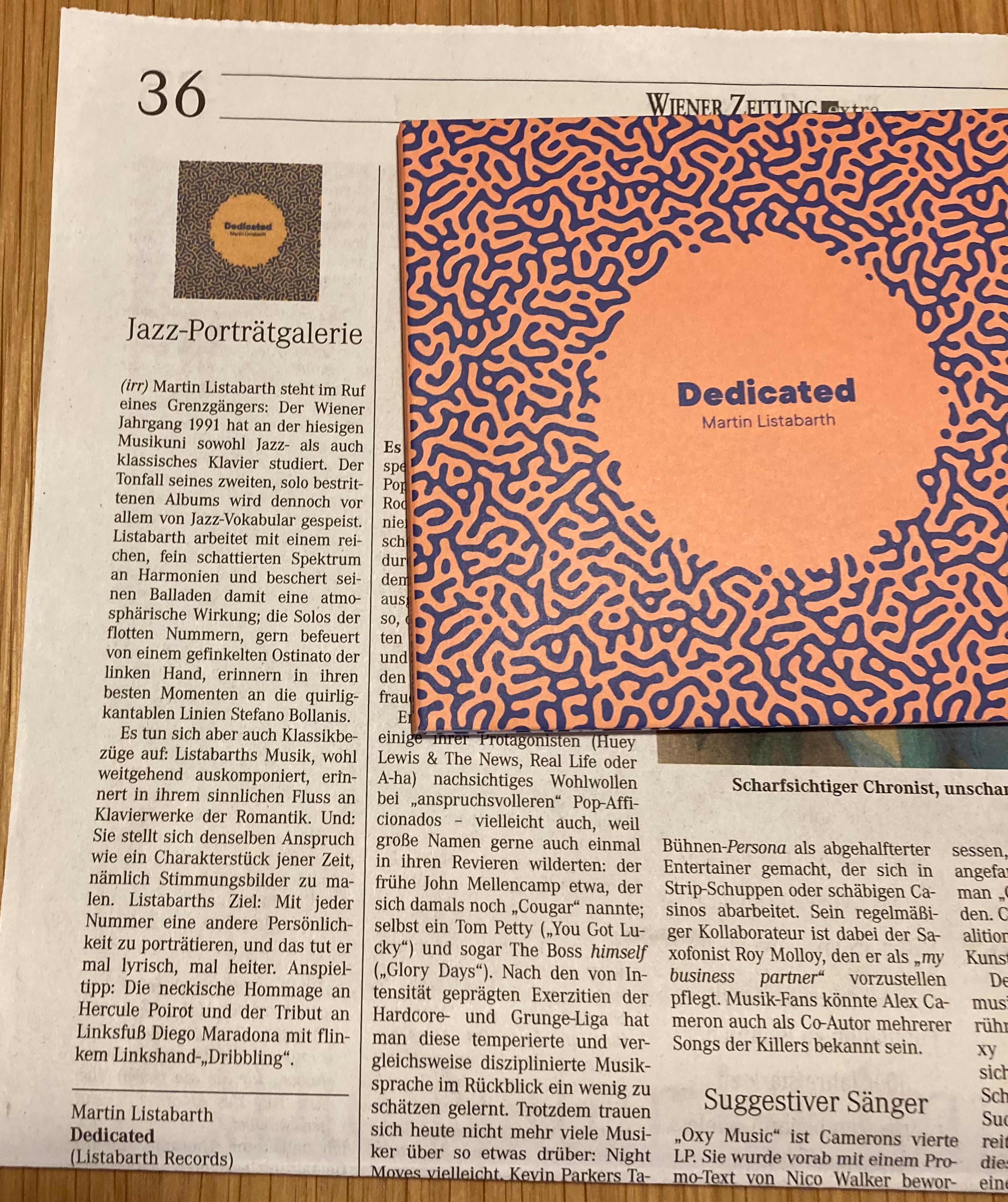Das Bild zeigt einen Artikel in der "Wiener Zeitung" über das neue Album "Dedicated" und die CD "Dedicated" auf einem Tisch liegend. 