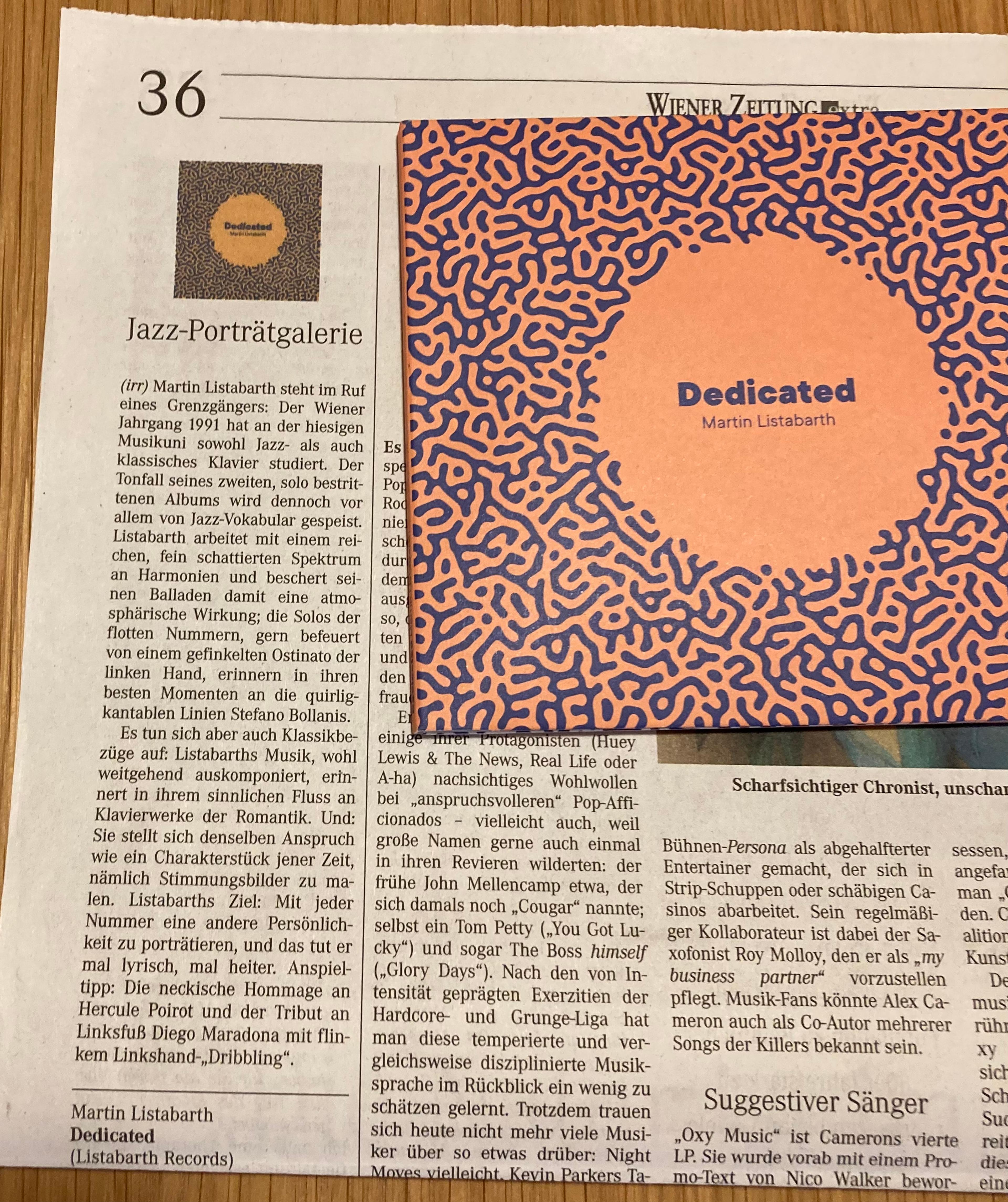 Das Bild zeigt einen Artikel in der "Wiener Zeitung" über das neue Album "Dedicated" und die CD "Dedicated" auf einem Tisch liegend.