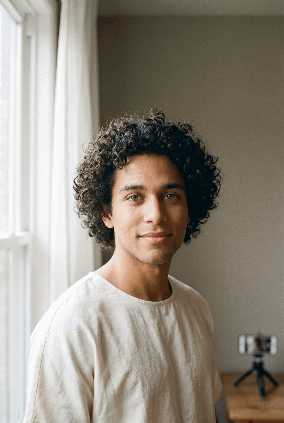A person standing near a large window with soft daylight falling on their face, neutral background, bright and natural lighting — smartphone on tripod.